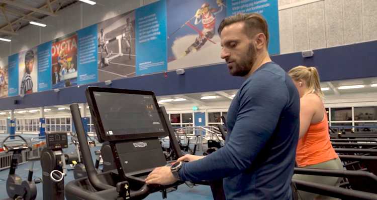 Inomhusmiljö, gym. En man med träningskläder står på ett löpband och håller sig i handtaget framför sig.