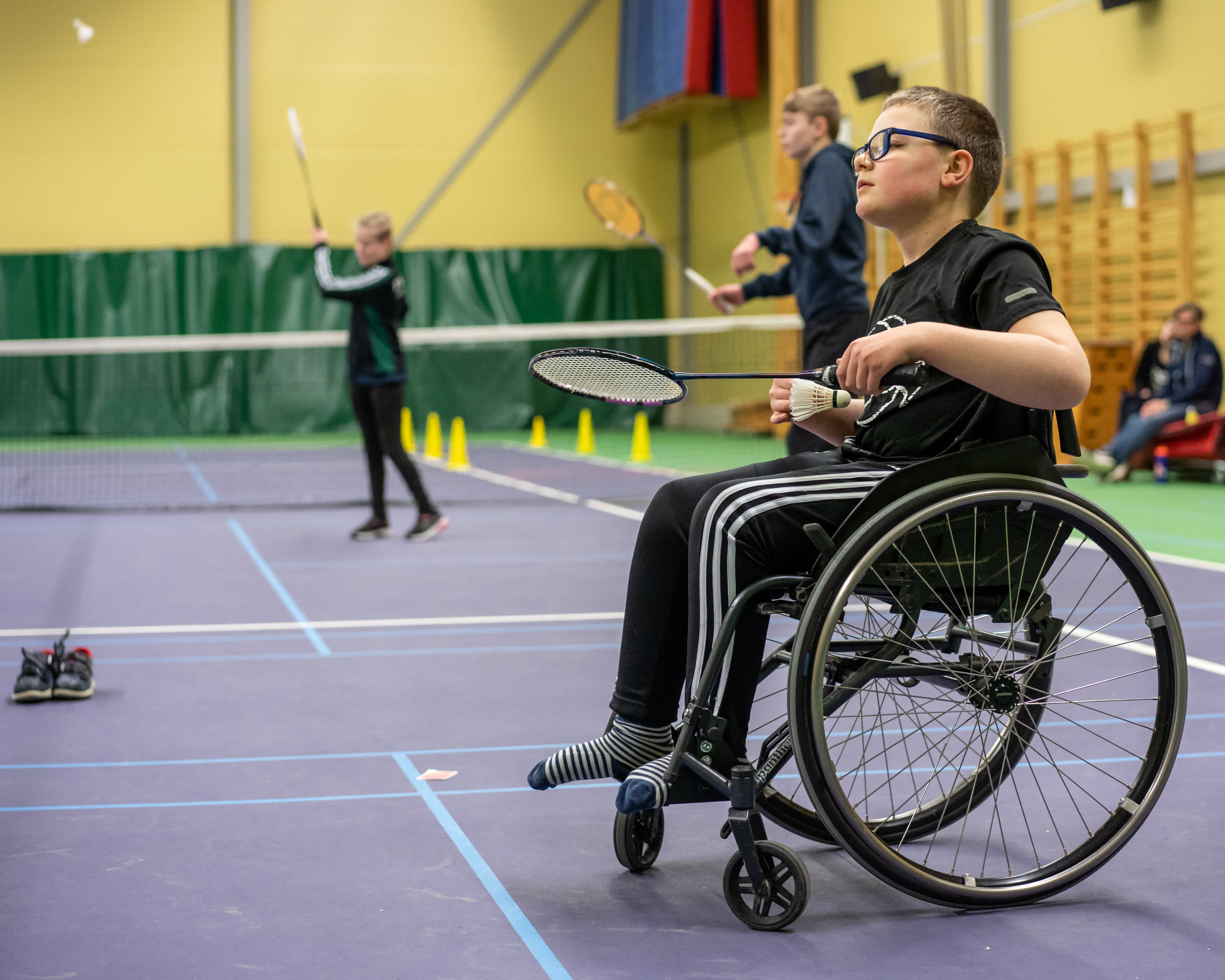 Pojke i rullstol gör sig redo att skjuta iväg badmintonfjädern. Flera badmintonspelare i bakgrunden.