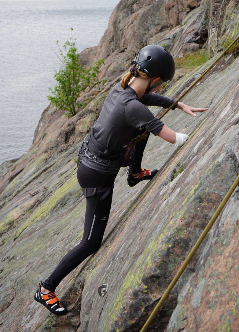 Utomhusmiljö, bergsvägg med vatten nedanför. Bild på en klättrare i profil, hon har hjälm och saknar en hand. Foto: Nacka Värmdö Klätterklubb.