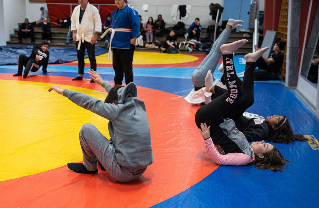 Teknikträning på judomattan, deltagarna övar på att rullar bakåt på ryggen. Mattan är en cirkel med gult i mitten, sedan en röd cirkel och längst ut blått.