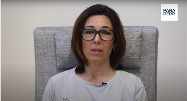 En kvinna sitter i en grå fåtölj. Hon har svarta glasögon, mörkbrunt axellångt hår och vit tröja. 