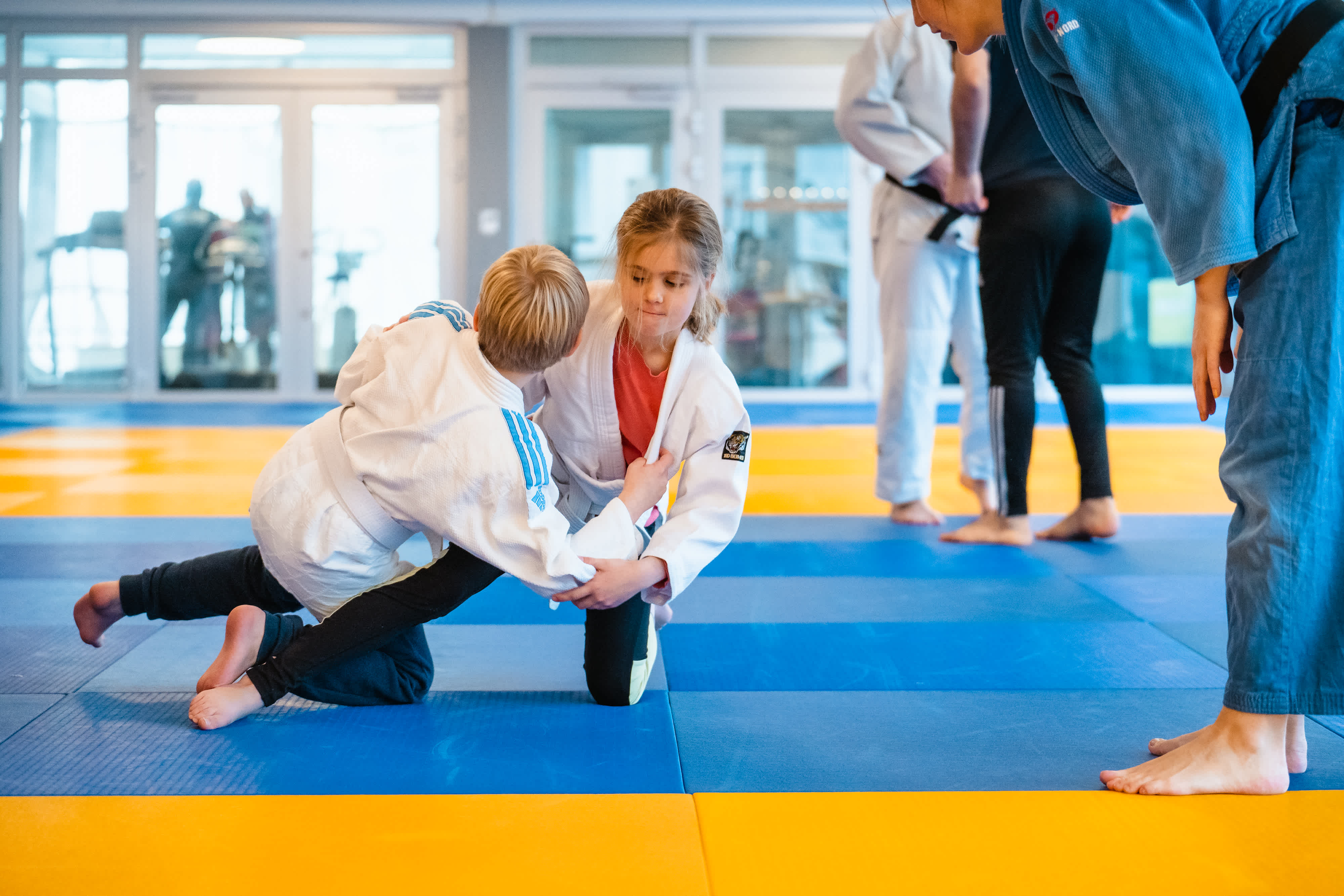 Inomhusmiljö, judomatta. Två barn i judodräkter testar ett judogrepp under uppsikt av en ledare.