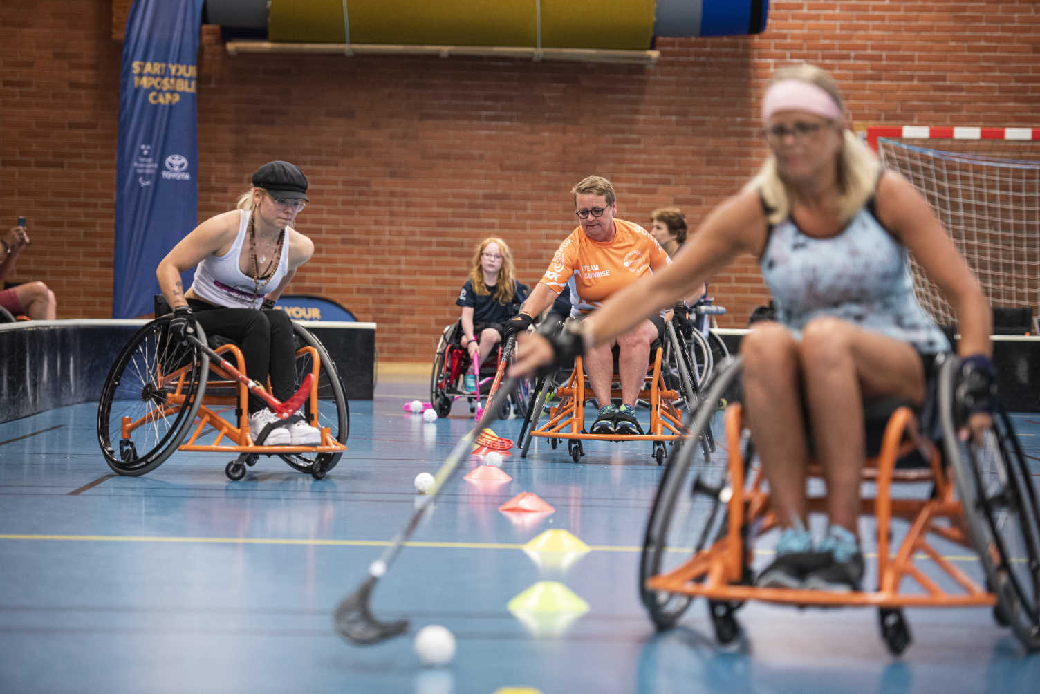 Tjejer i olika åldrar tränar rullstolsinnebandy mellan koner i idrottshall.