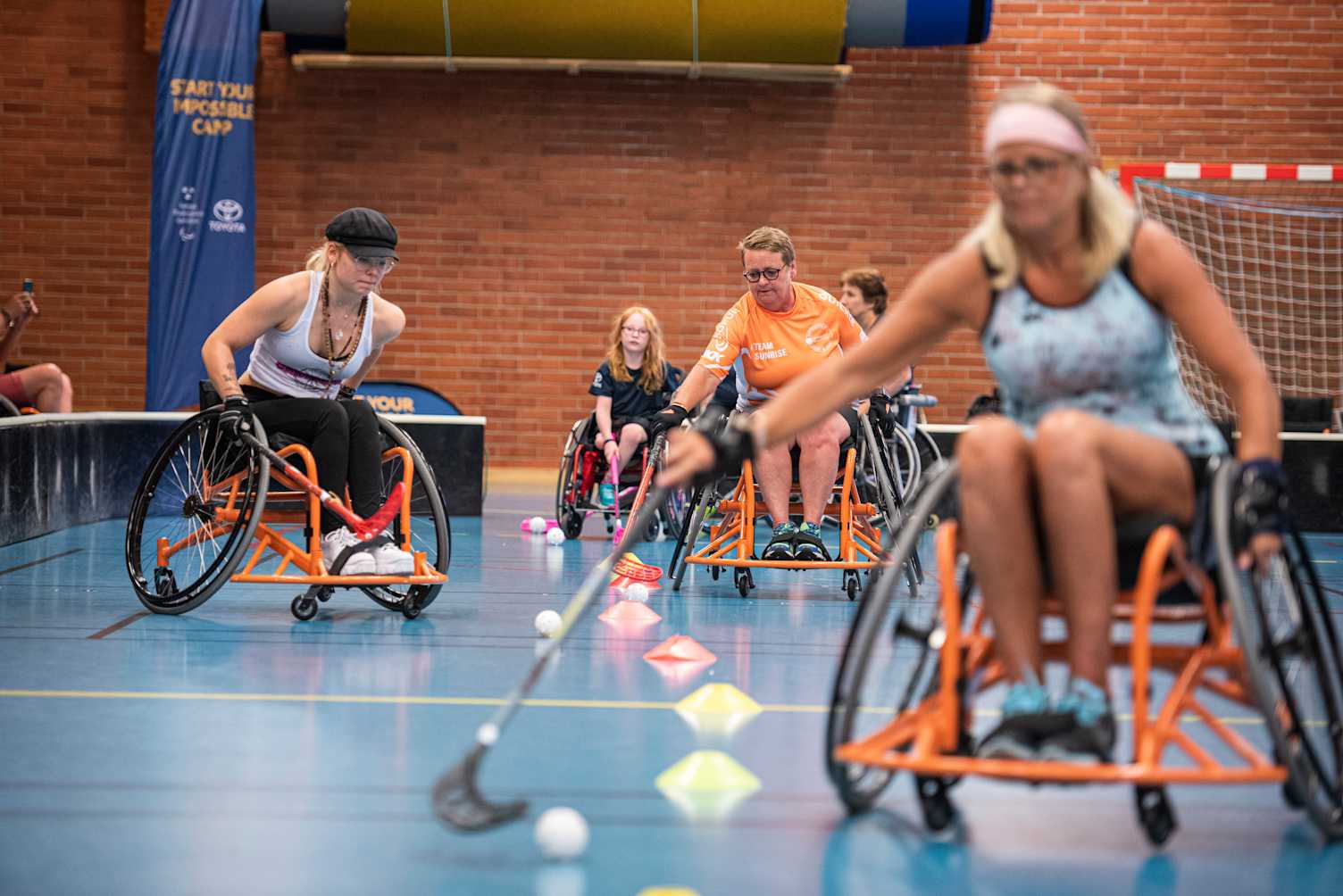 Tjejer i olika åldrar tränar rullstolsinnebandy mellan koner i idrottshall.