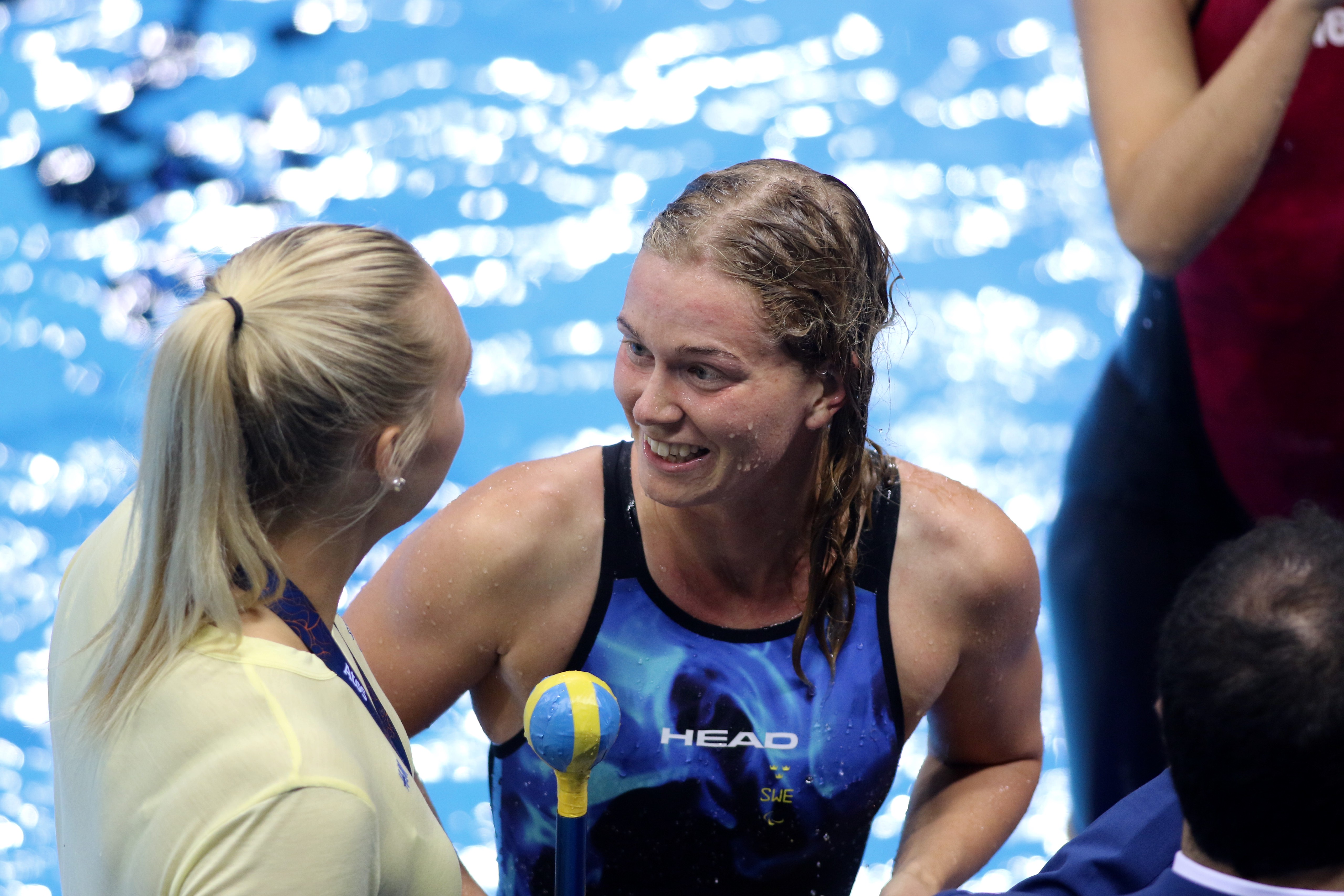 Simmaren Maja Reichard står bredvid bassängen med sin teamkollega. Kollegan håller i en pinne med en boll i ena änden.