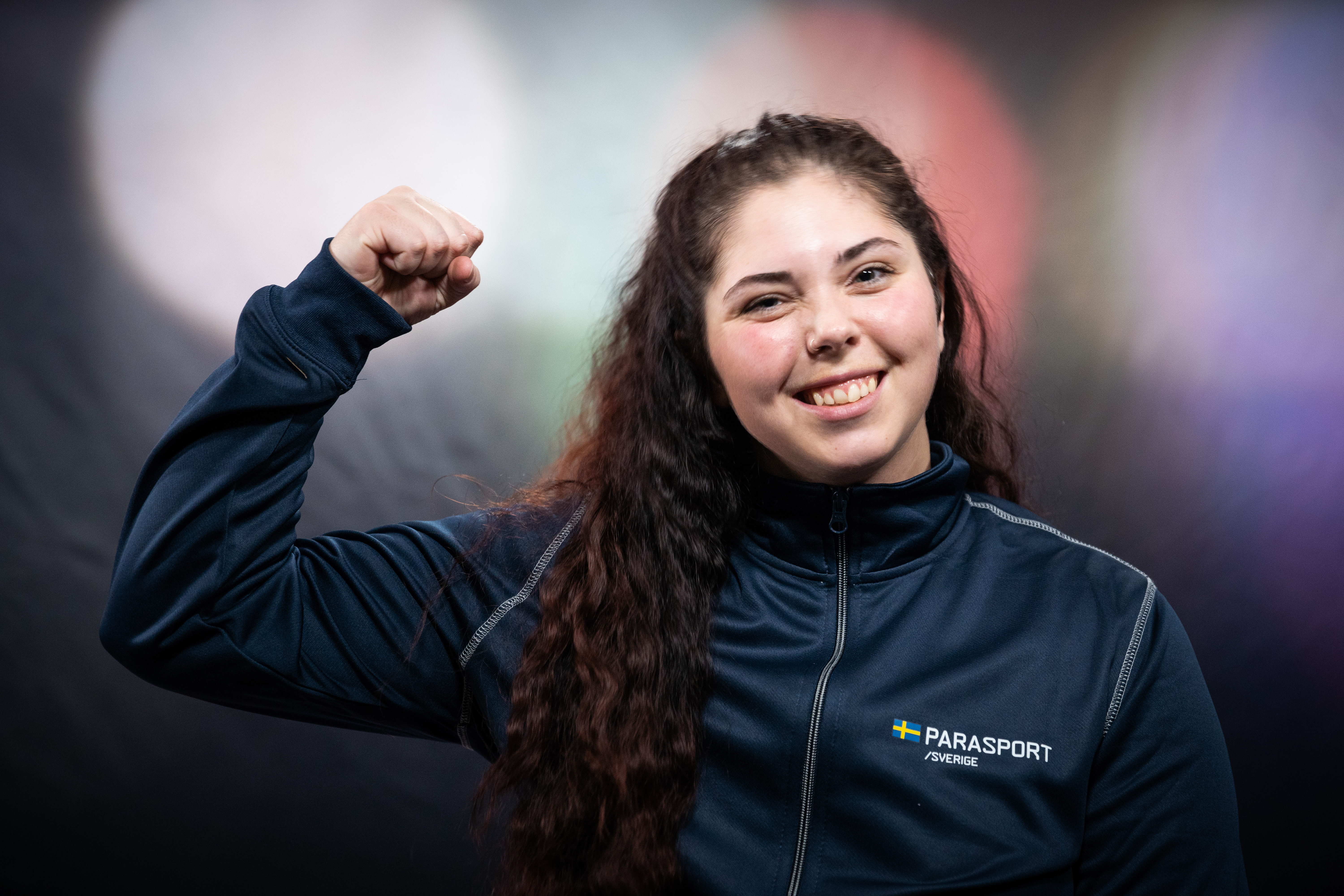 Kvinna med långt, mörkt och uppsatt hår. Håller upp armen, böjer armbågen, har handen i en knytnäve. Hon visar att hon är stark. Kvinnan har ett stort leende, blinkar med ena ögat. På sig har hon en blå tröja med texten Parasport Sverige på. I bakgrunden syns cirklar i olika färger.