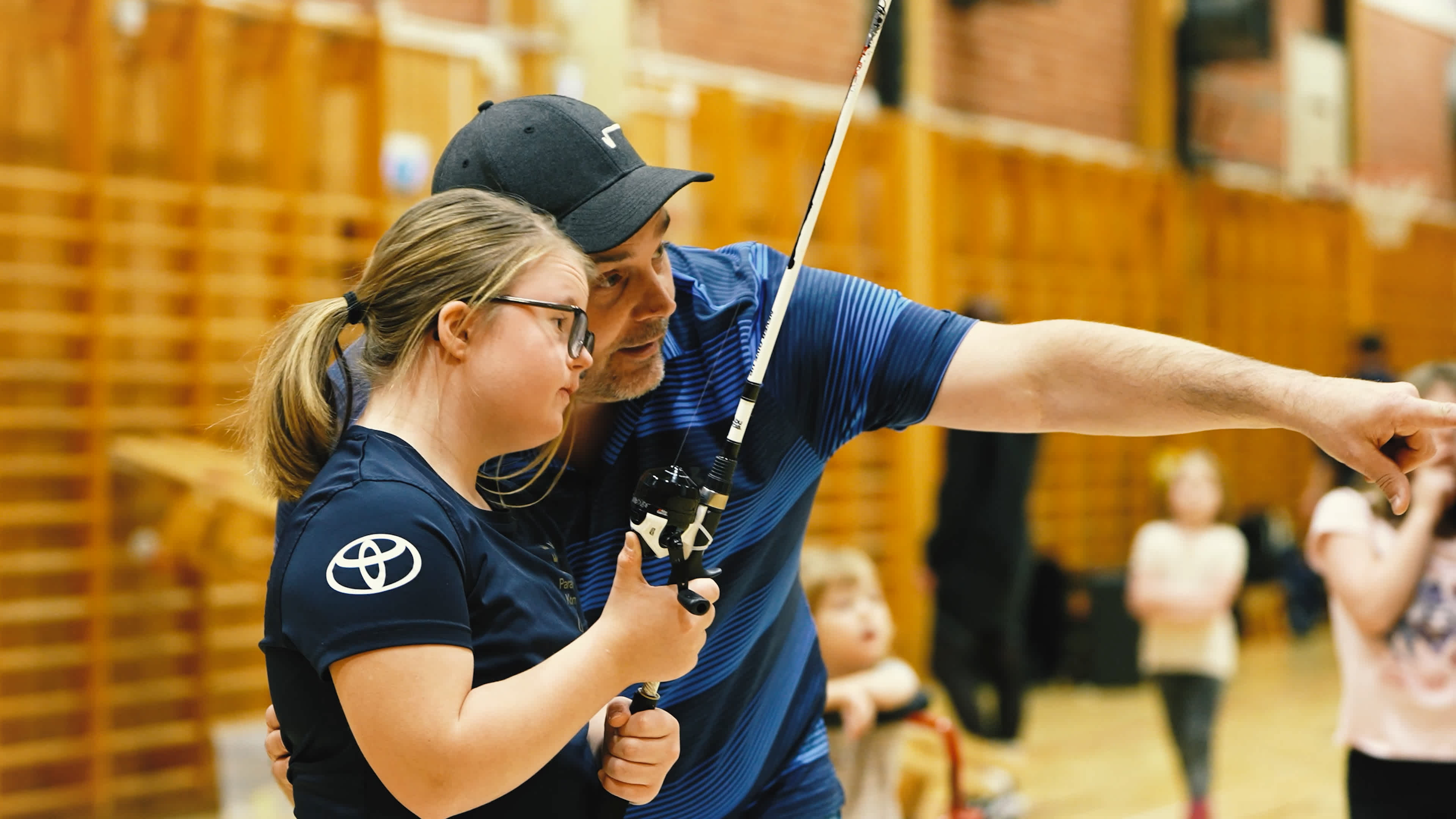 Inomhusmiljö. idrottshall. En ung tjej med ett castingspö i händerna får instruktioner av en ledare som pekar var hon ska sikta.