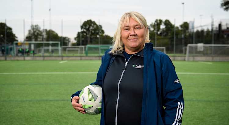 Utomhusmiljö, fotbollsplan. Charlotte, kvinna med blont hår och träningsjacka, står med en fotboll under armen.