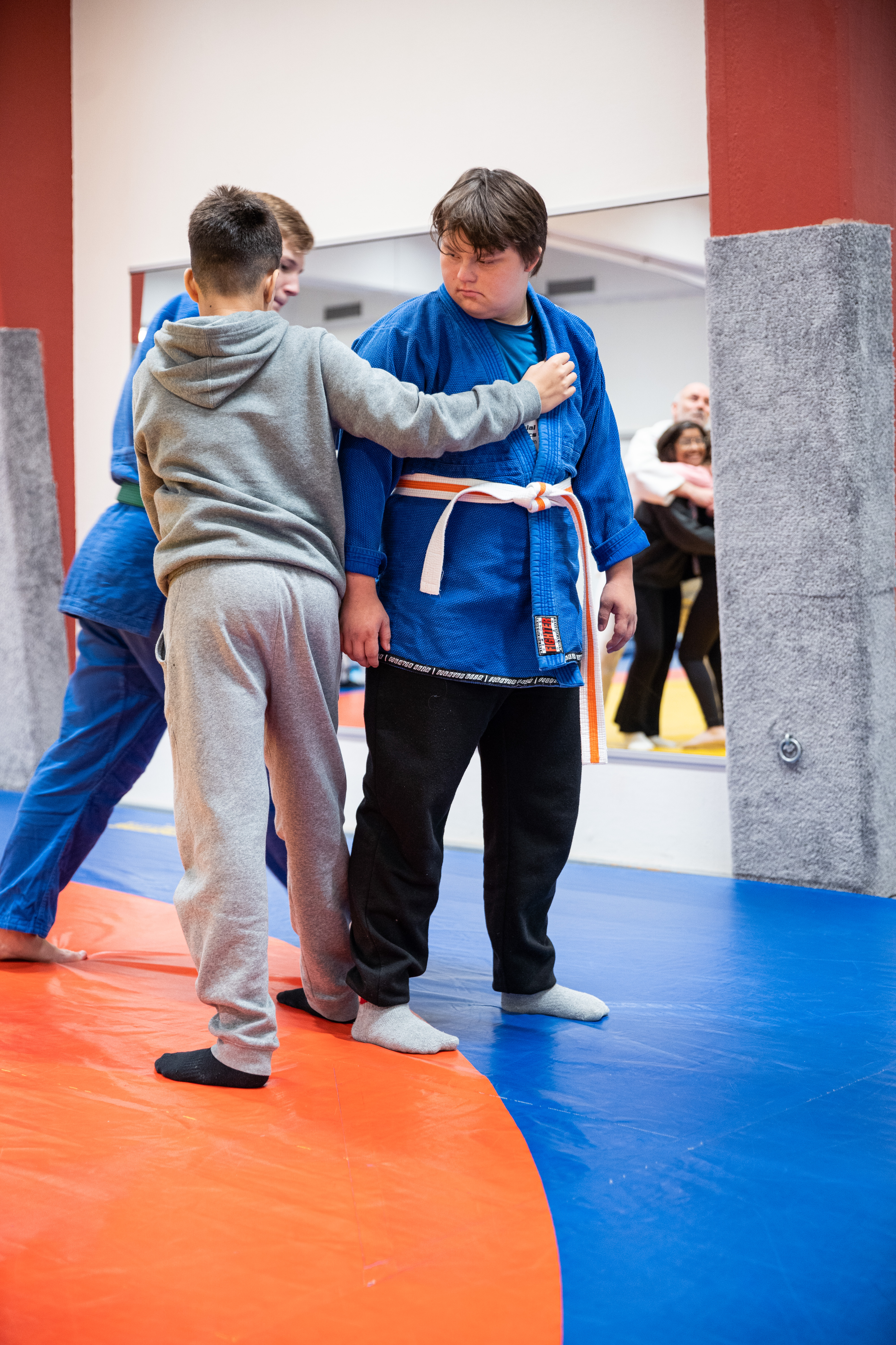 Två personer på en judomatta går tillsammans med instruktör igenom hur man fäller varandra i judo. Mattan är en cirkel med gult i mitten, sedan en röd cirkel och längst ut blått.