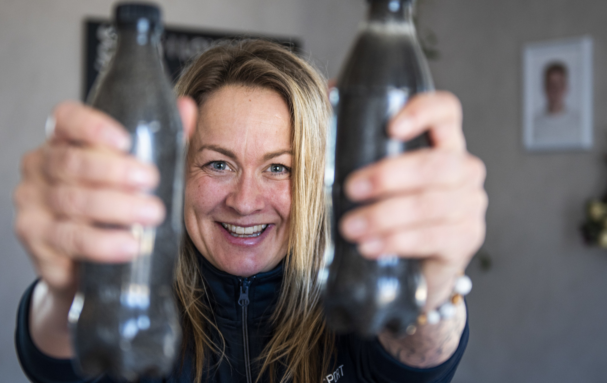 En kvinna med ljust hår och som ler stort, håller upp två sandfyllda petflaskor framför sig. 