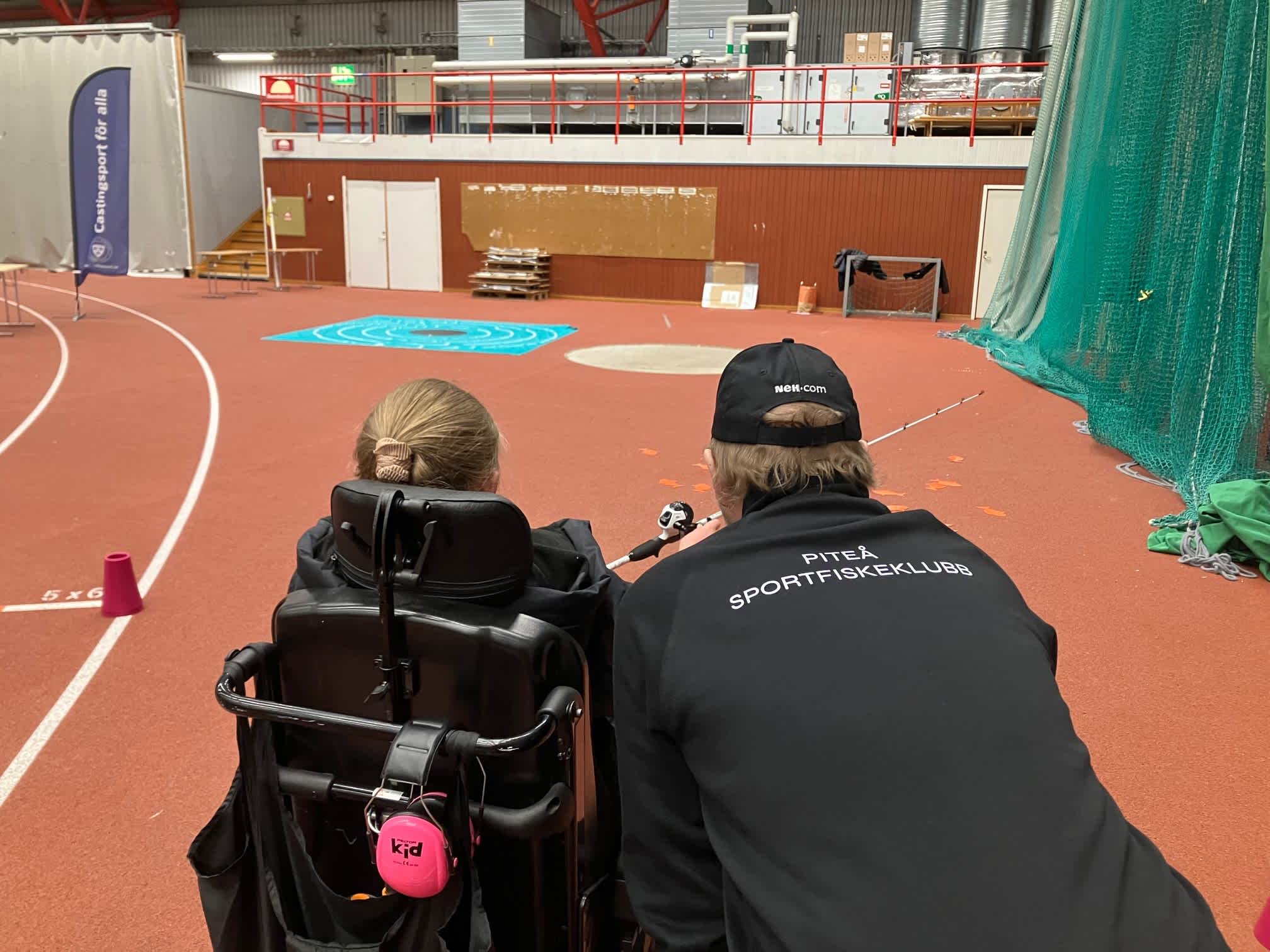 Inomhusmiljö, idrottshall. Ryggarna på en deltagare i rullstol och en ledare. I bakgrunden syns den stora tavlan liggande på golvet, som deltagaren ska pricka.
