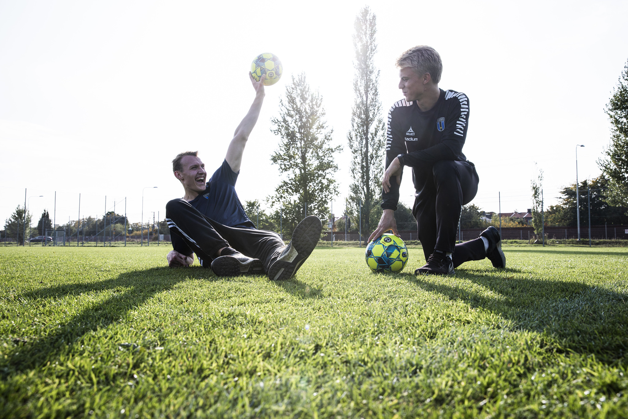 Två män är på en gräsmatta. Den ena ligger på ryggen och stöttar upp överkroppen med ena armen, och håller en fotboll upp i luften med andra armen med ett stort leende. Den andra står på ett knä och håller armbågen på det andra knäet. Han håller handen på en fotboll medan han tittar på den första mannen.