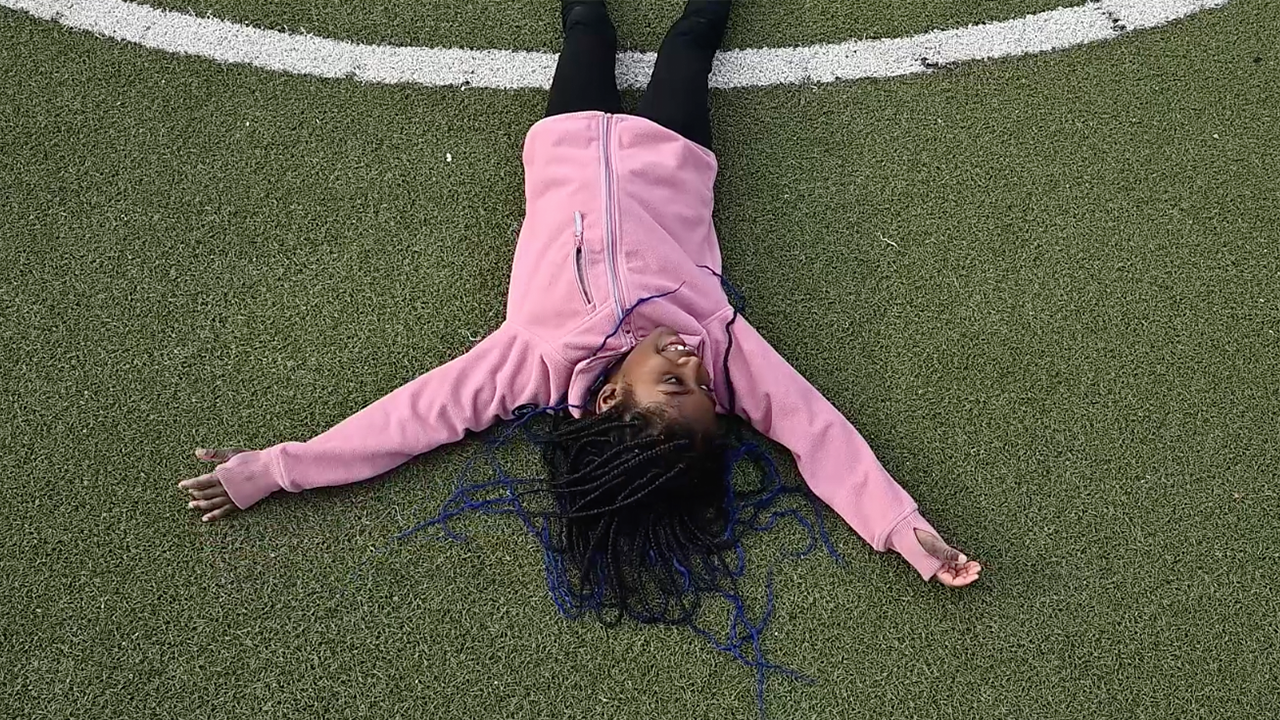 Utomhusmiljö. Foto taget ovanifrån. Alicia som ligger på rygg på fotbollsplanen med utsträckta armar.