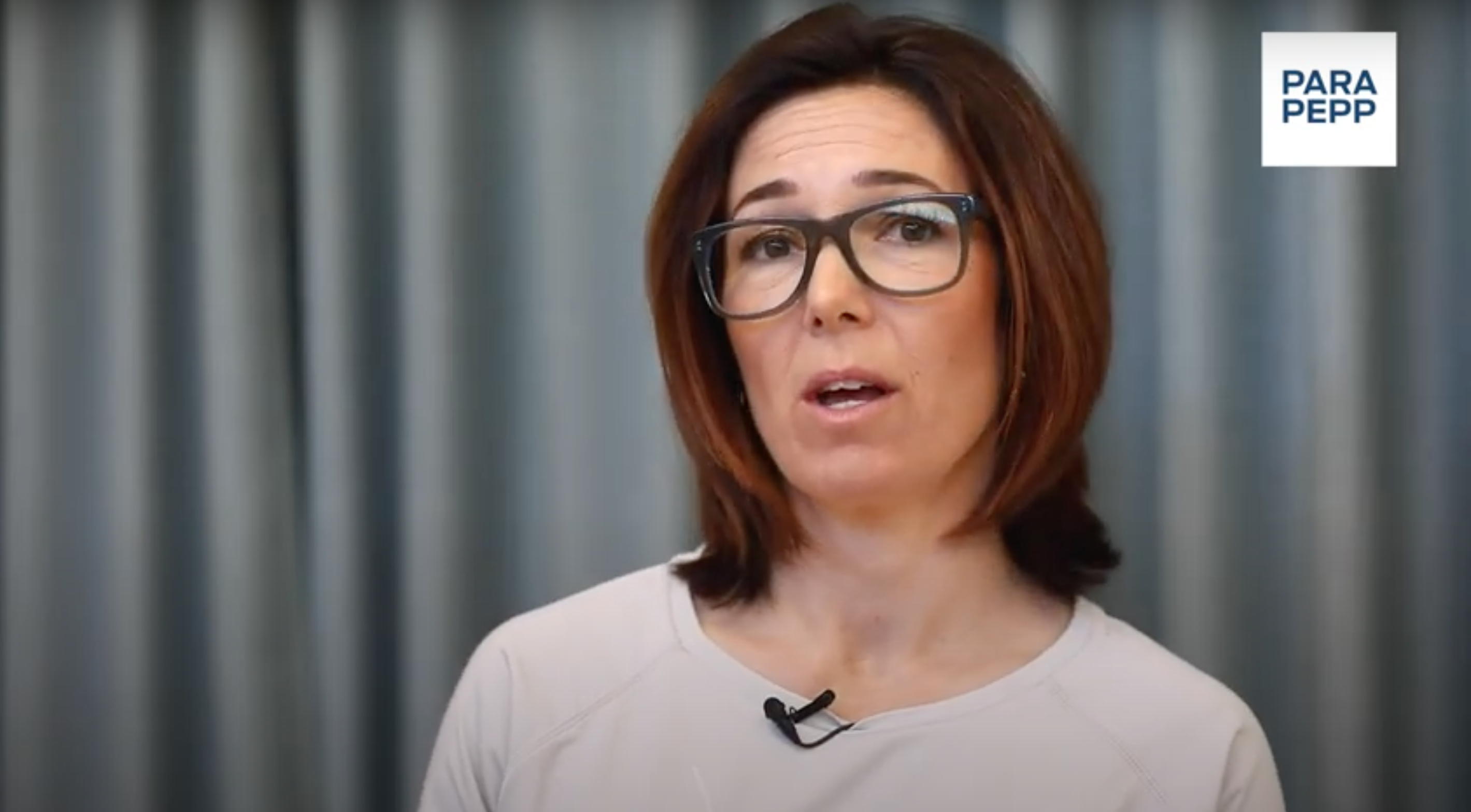 En kvinna med axellångt, mörkbrunt hår och svarta glasögon pratar in i kameran. Hon har ett allvarligt ansiktsuttryck, hennes tröja är ljus och bakom henne är ett grått draperi. 