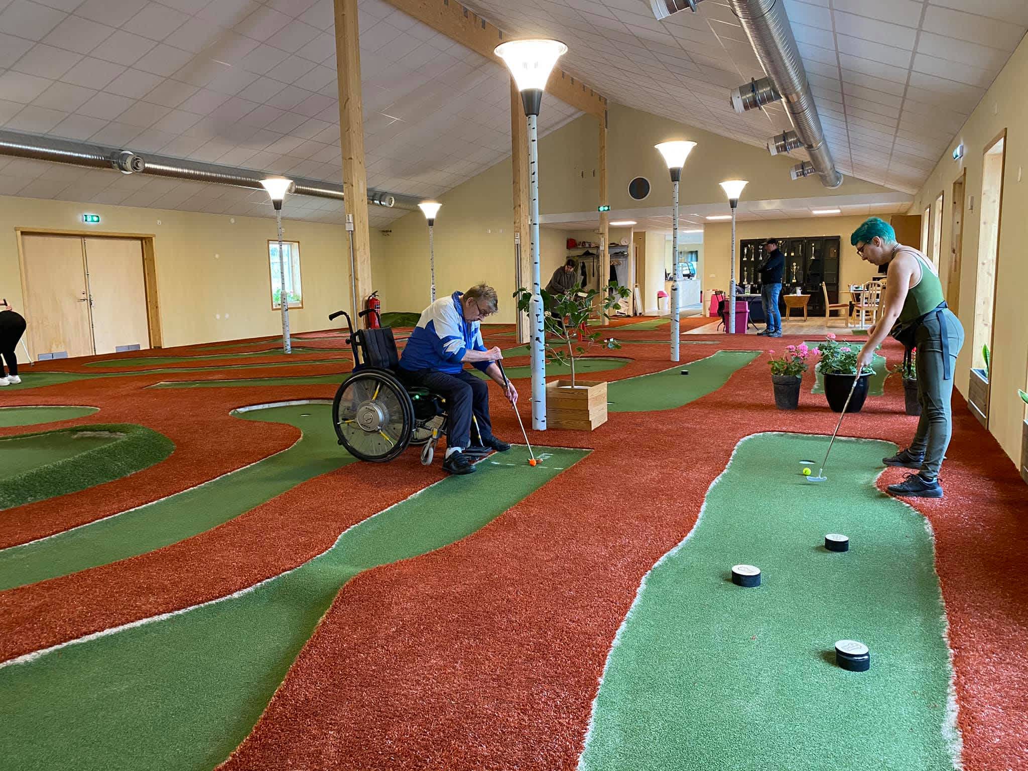 En person sitter i rullstol och ska putta iväg golfbollen. En annan person står upp och ska putta iväg bollen.