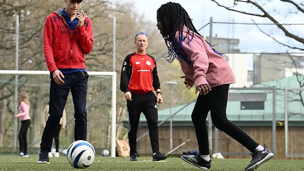 Utomhusmiljö, fotbollsplan. Foto på Alicia, en ung flicka som sparkar fotbollen framför sig. I bakgrunden lagkamrat och tränare.