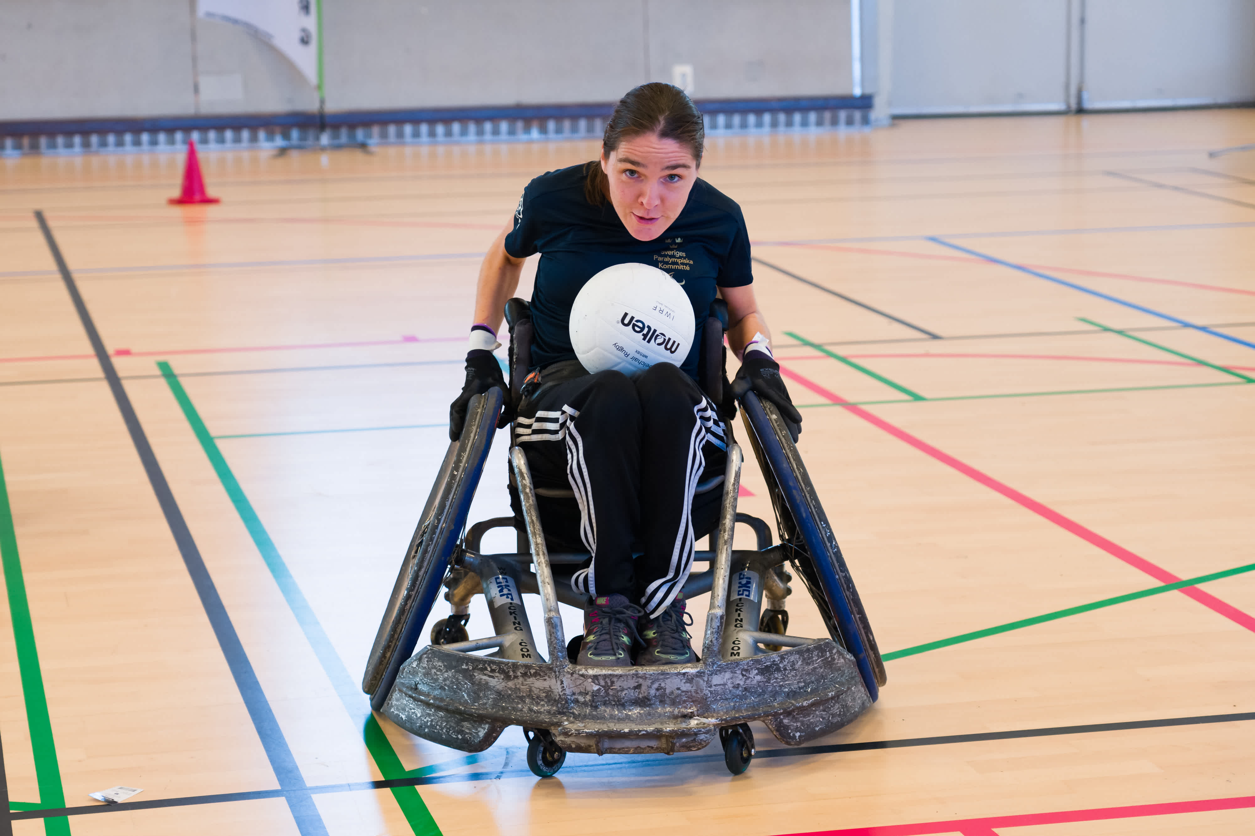 Ung kvinna i rugbystol med rugbyboll i knät kör målmedvetet mot kameran.