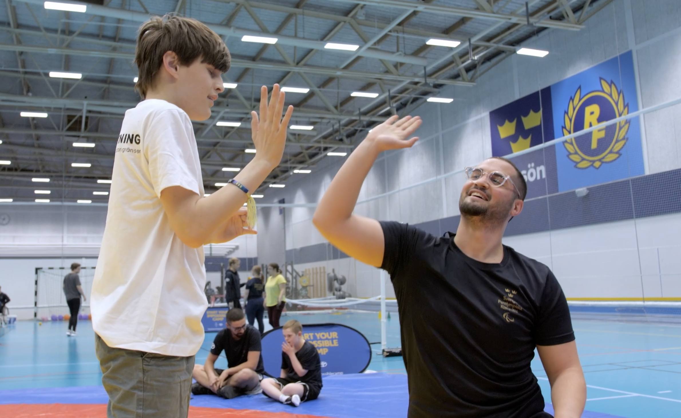 Brottningsledaren Cem gör high five med deltagaren Adam, som precis har provat på brottning. De är i en upplyst idrottshall med blå golv, de står på en brottarmatta.