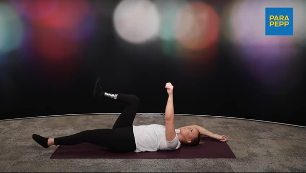 En kvinna ligger på rygg på en träningsmatta med vänster ben och vänster arm i luften. På vänster underben har kvinnan en protes. Hon har vit t-shirt och svarta träningsbyxor. Bakgrunden är mörk med stora, suddiga cirklar i olika färger.