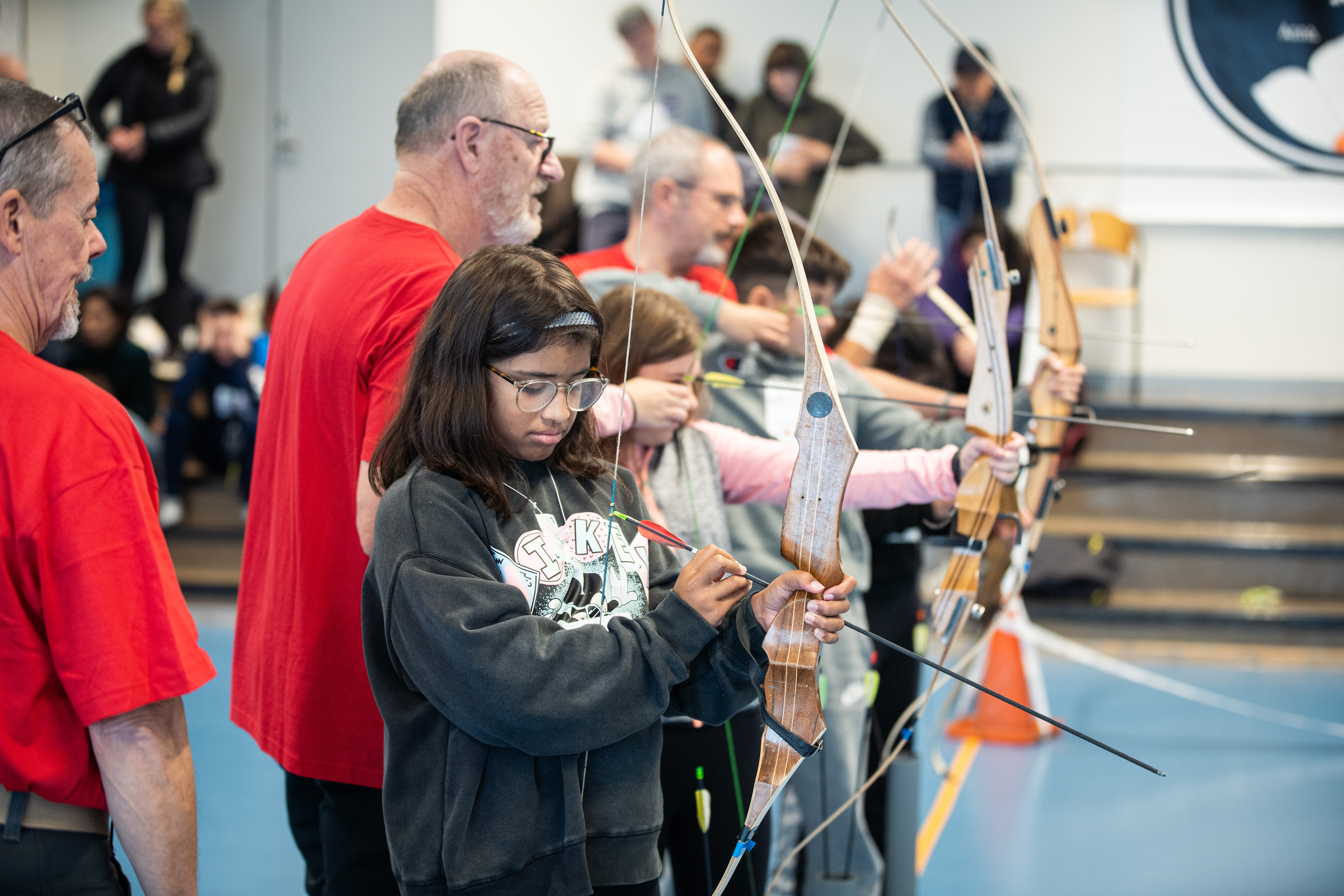 Flicka laddar sin båge med en pil. Flera skyttar i bakgrunden.