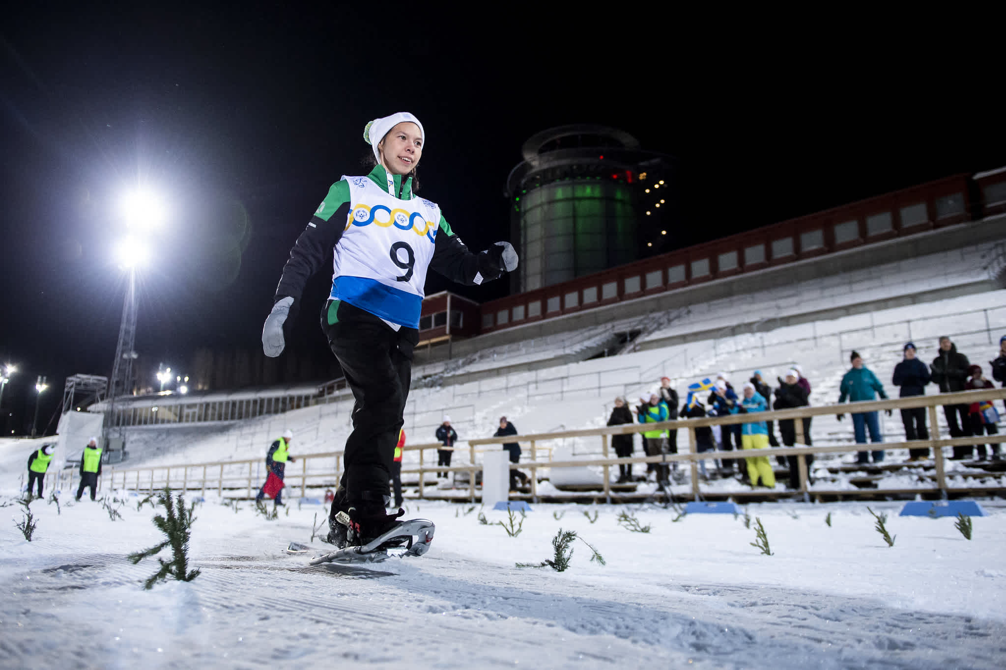 Tävlande i snöskolöpning på Östersund skidstadion. På läktaren står publik som hejar på deltagarna.