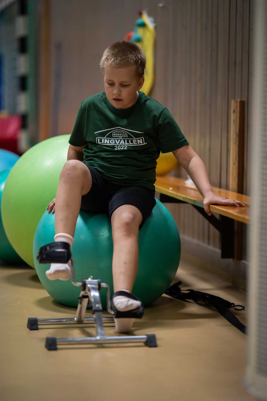 Pojke tränar benen på cykelmaskin, sittandes på en pilatesboll. 