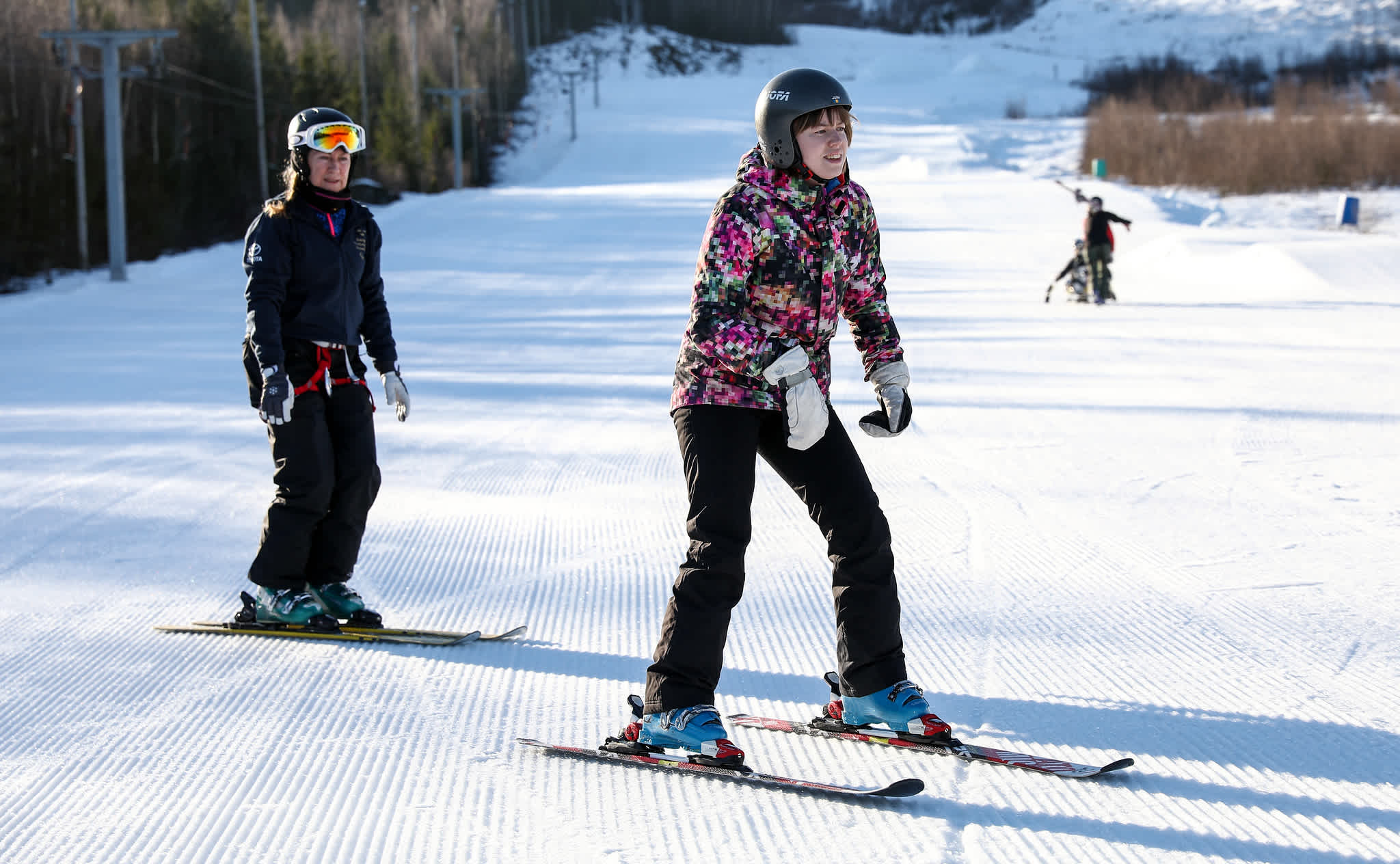 En tjej på skidor åker slalom på en flackare del av backen. Med henne åker en lärare.