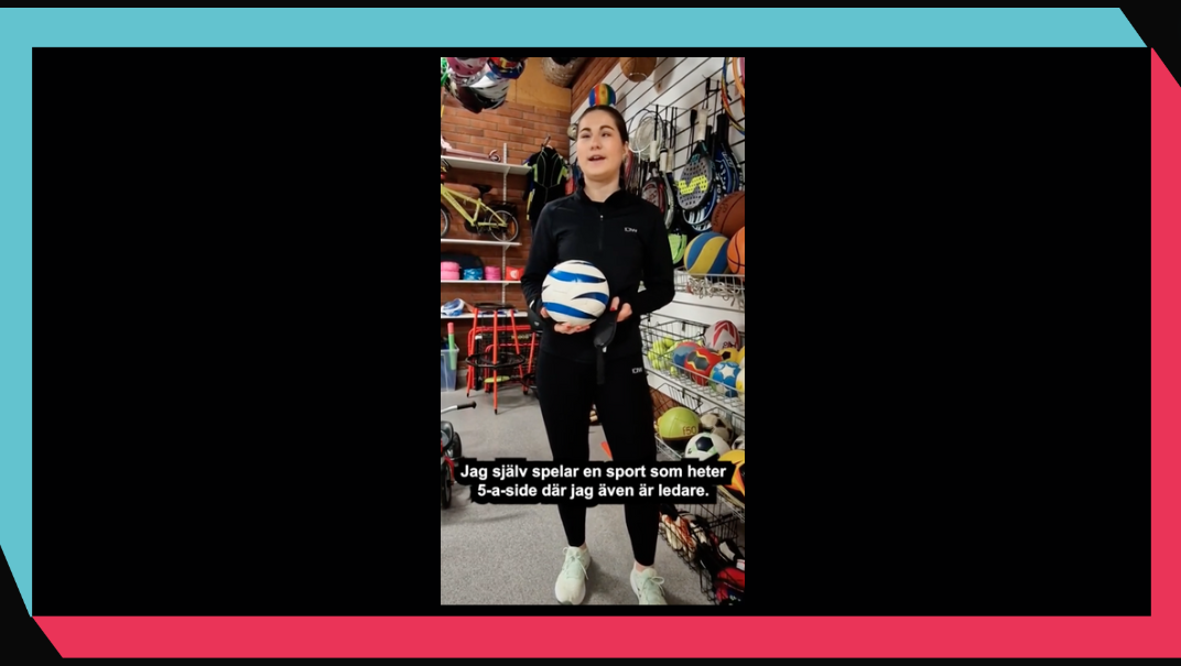 Inomhusmiljö, butik. En ung kvinna står framför en vägg med sportutrustning. I handen har hon en blåvit fotboll.