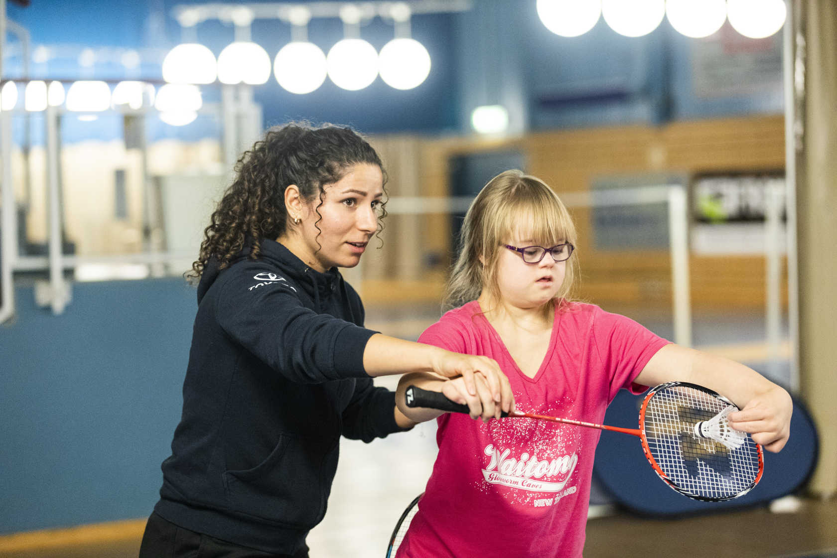 En tjej får hjälp av en ledare som visar hur hon träffar badmintonfjädern med racket.