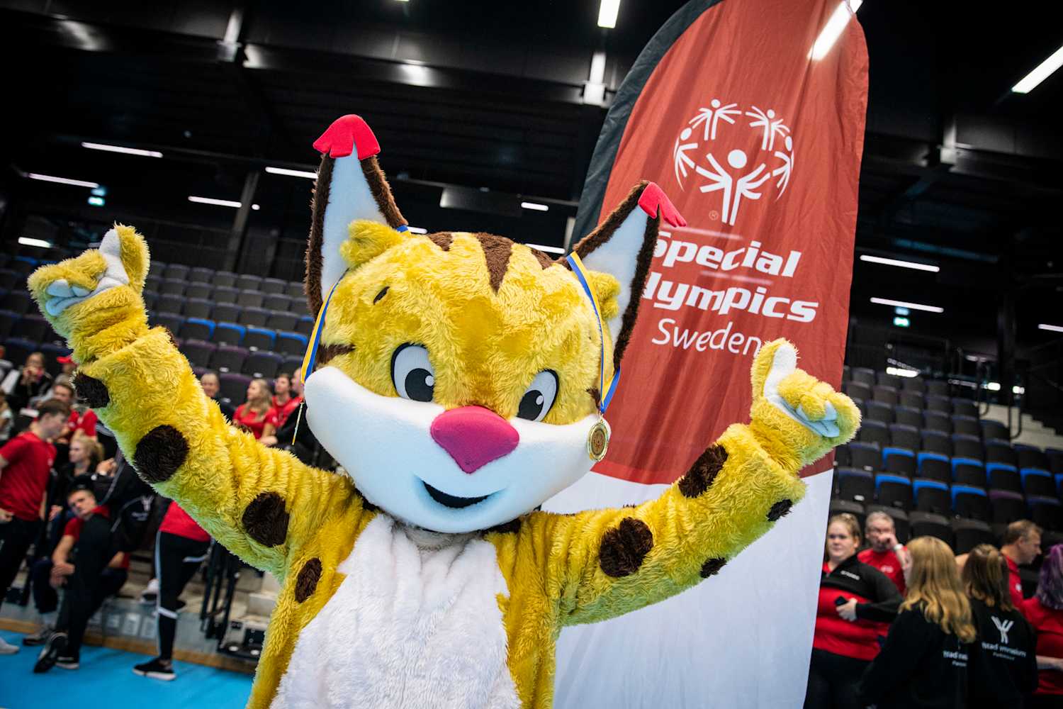 Inomhusmiljö, idrottshall. Maskoten Speci-Lo är ett lodjur, hon håller upp händerna framför en Special Olympics-flagga.