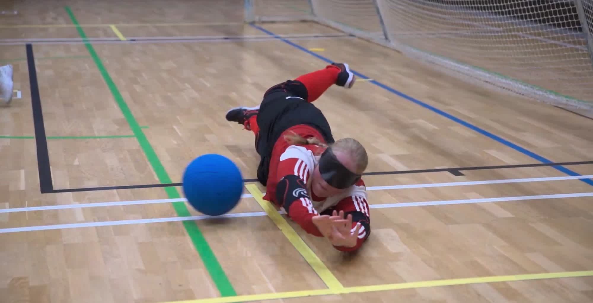 Inomhusmiljö, idrottshall. En goalballspelare med träningskläder och ögonbindel kastar sig längs golvet för att skydda målet från den blå bollen som kommer farande.