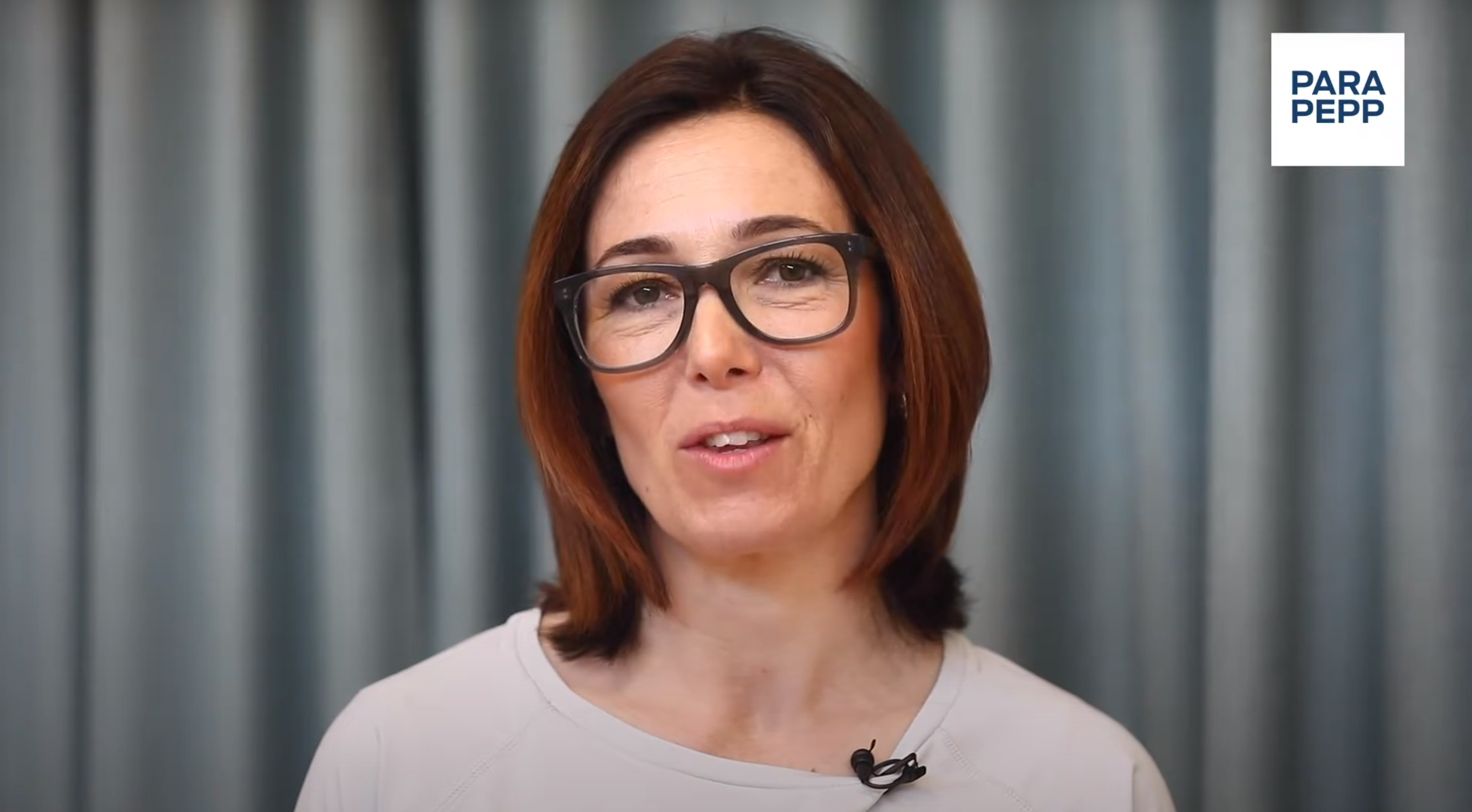 En kvinna med brunt, axellångt hår och svarta glasögon pratar leende in i kameran. Hon har en vit överdel och  bakgrunden är grå. 