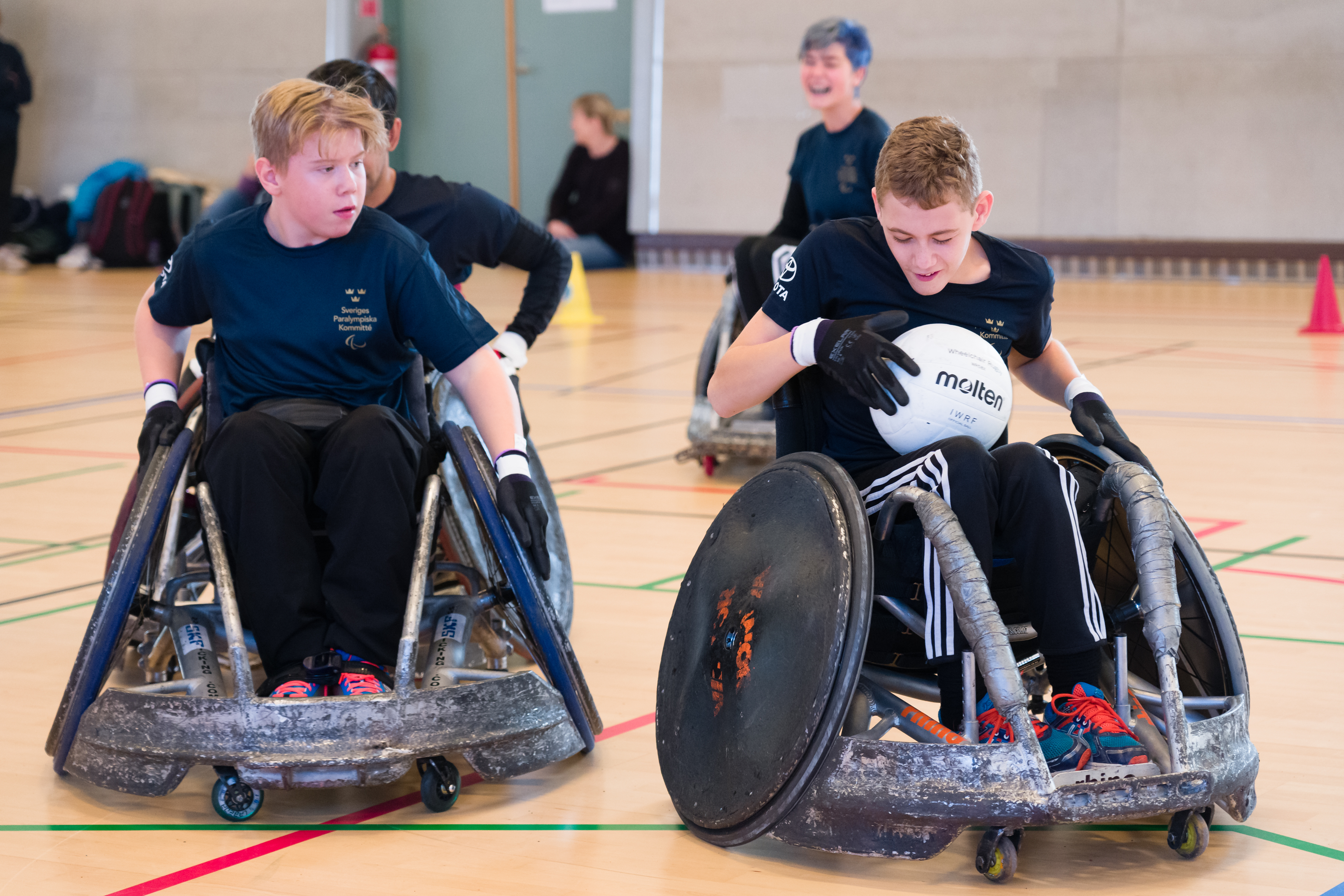 Två killar kör rullstolsrugby, den ena har bollen i knät. 