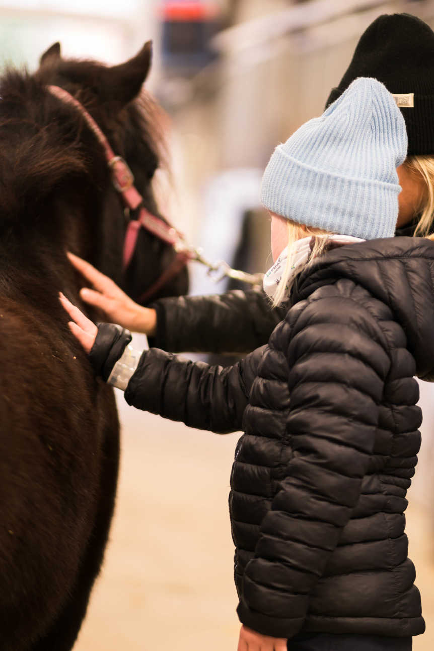 En ung tjej klappar en häst på magen. Hon har vinterkläder och mössa.