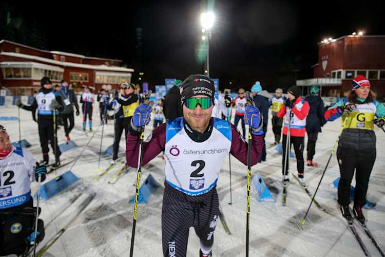 Åkare i Legacyloppet står uppradade och väntar på start. I mitten och fokus av bilden är en åkare med skidglasögon, redo med stavarna. Det är mörkt ute, strålkastare lyser upp stadion.