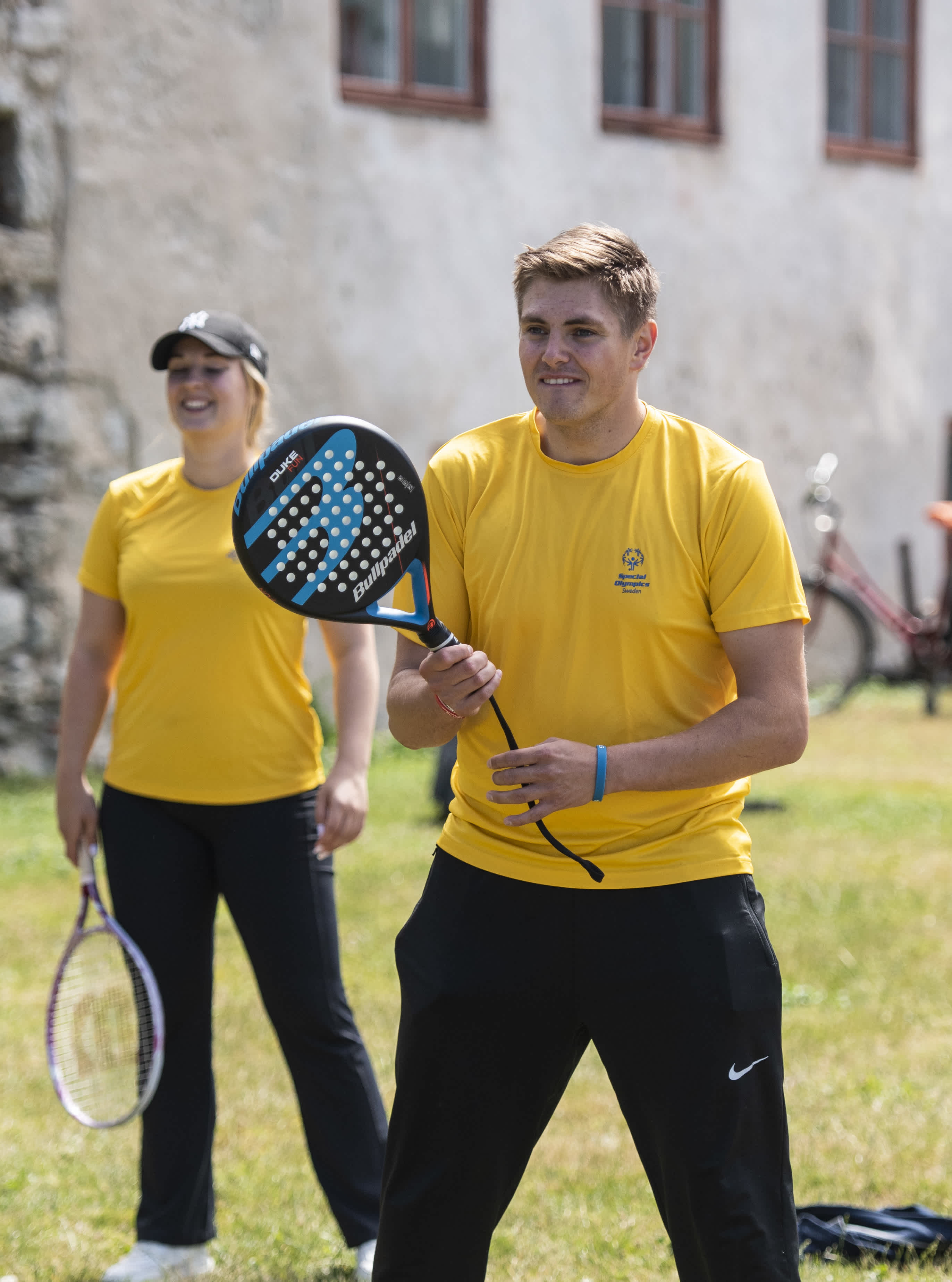 En man och en kvinna i gula tröjor spelar i samma lag. Han står redo med padelracket i höger hand.