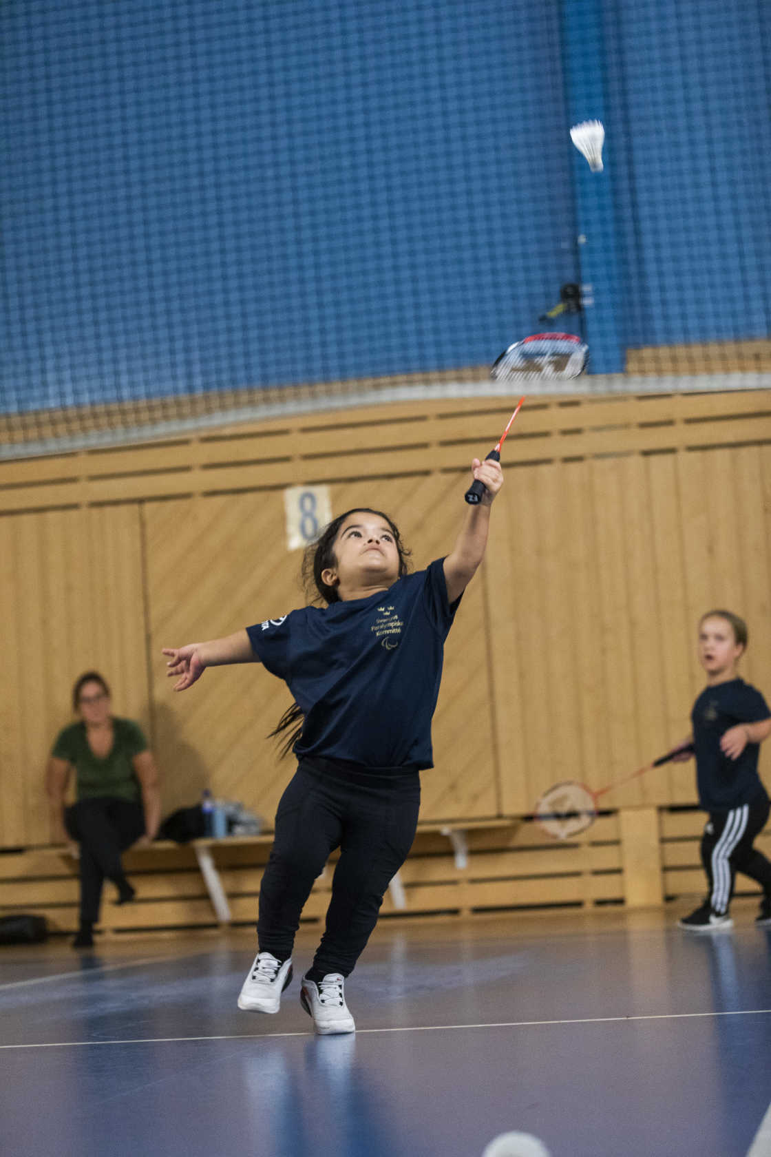 En tjej med kortväxthet sträcker sig för att nå badmintonfjädern. Hennes medspelare står bakom. 