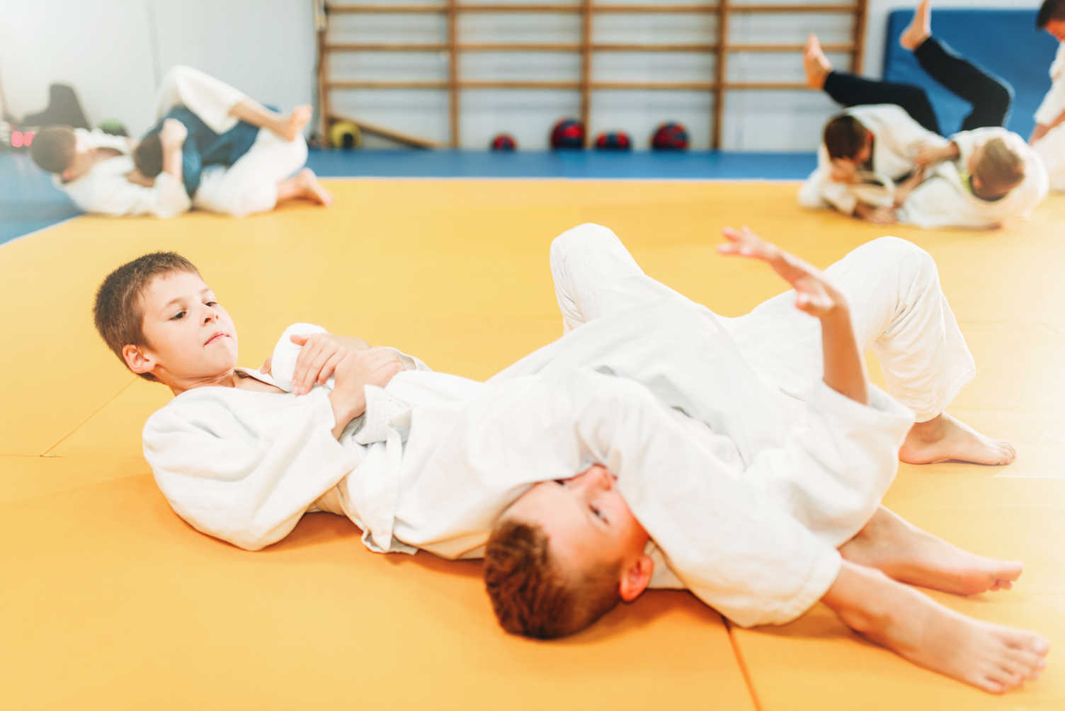 Två pojkar i kampsportsdräkt övar fasthållningsteknik liggandes på mattan. 