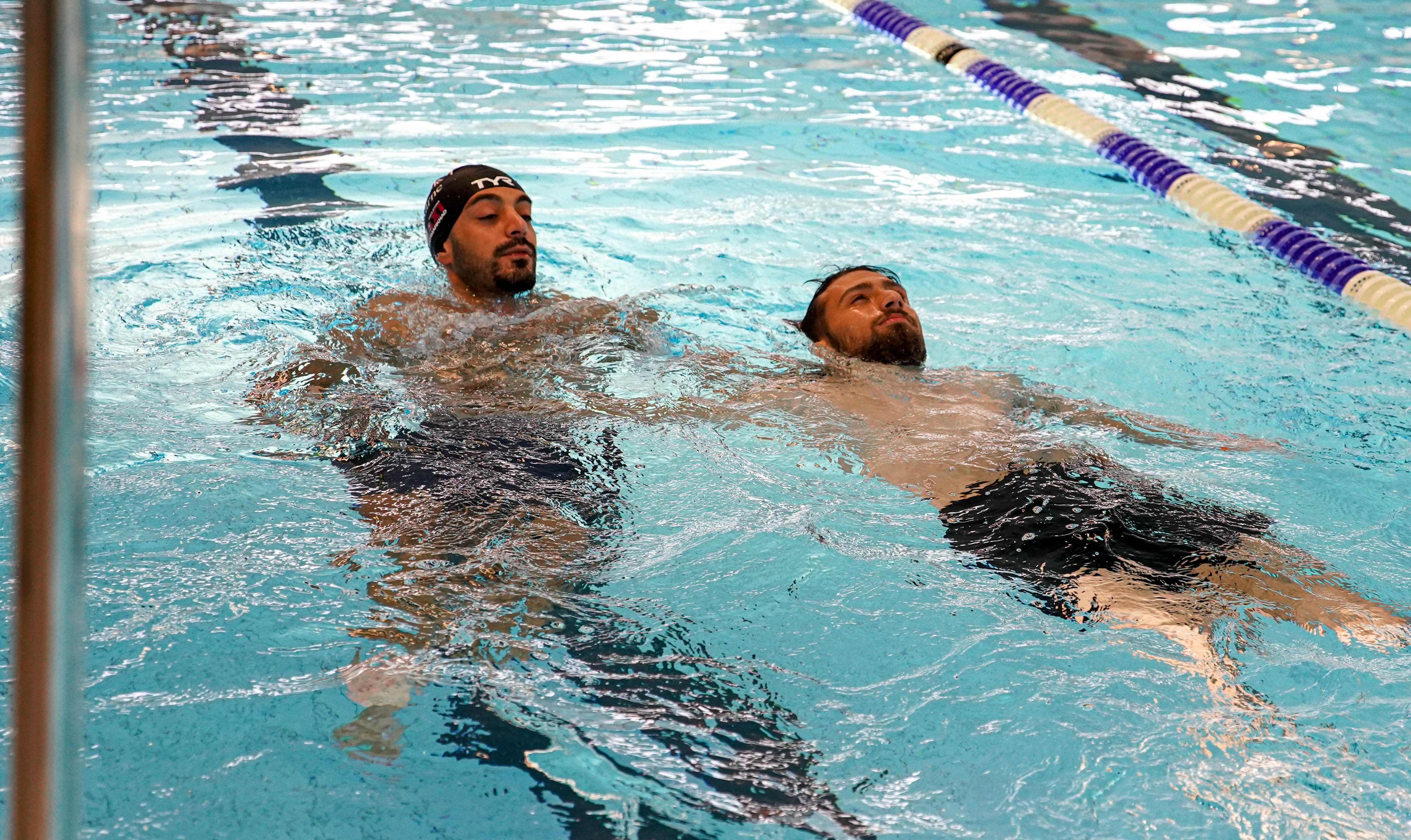 Två män tränar ryggsim tillsammans i bassäng inomhus.