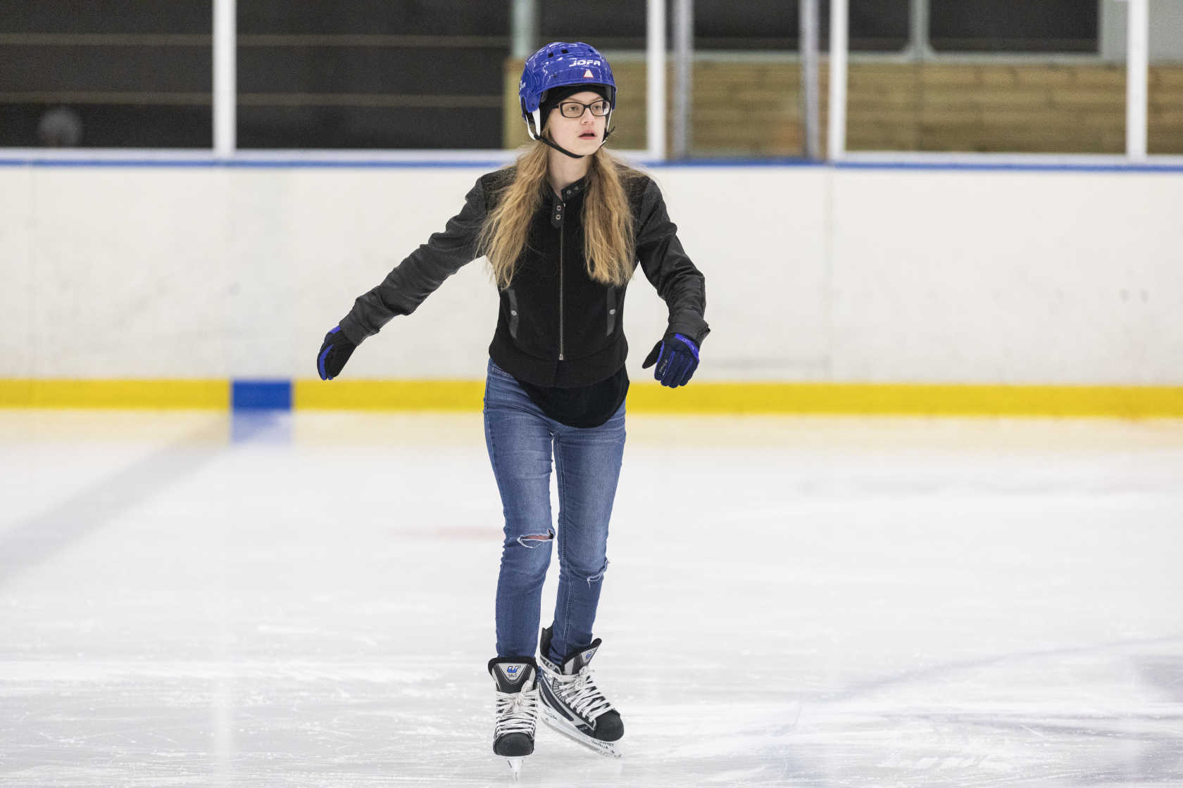 En tjej med blå skridskohjälp och hockeyskridskor åker på en rink i en ishall.
