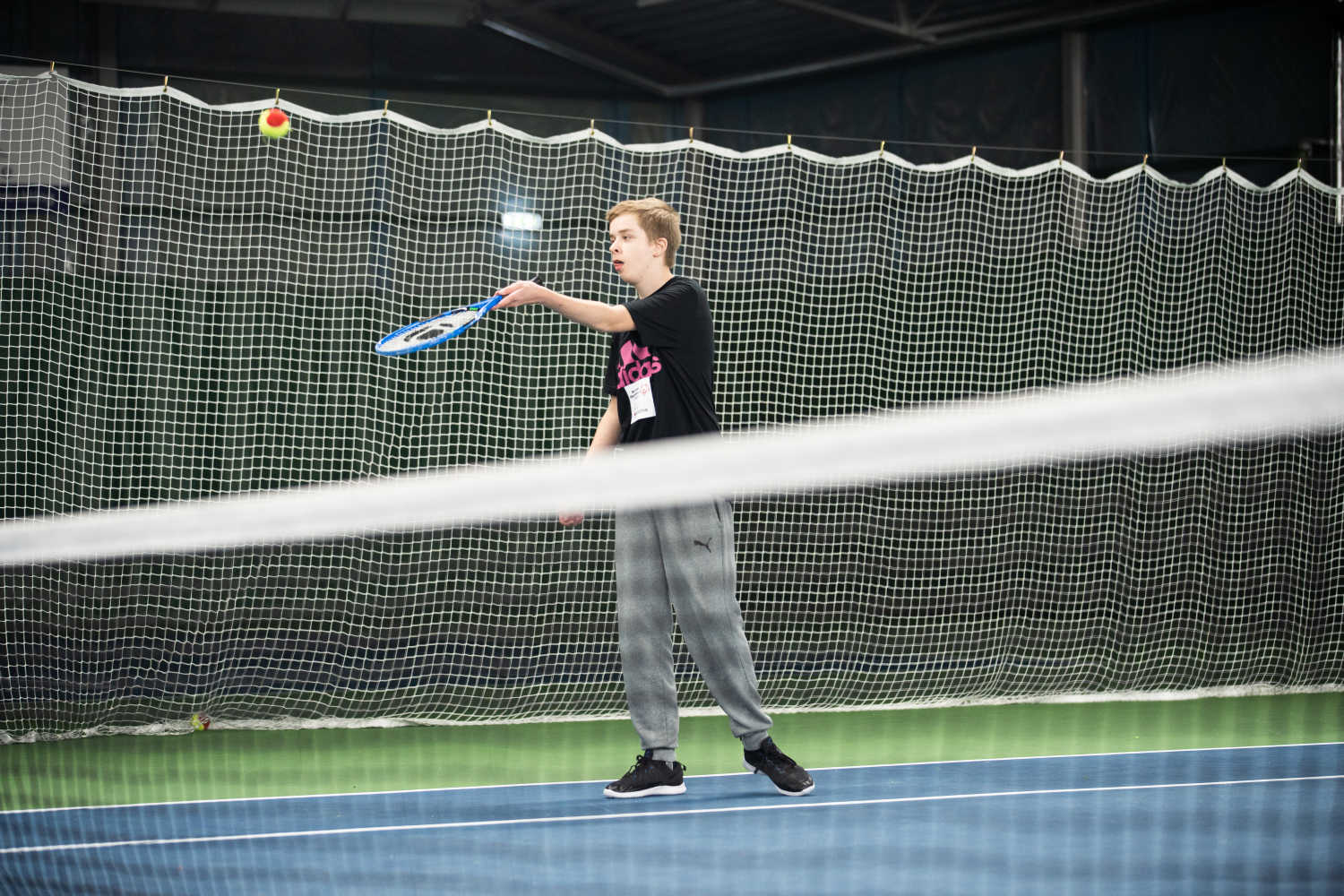 Ung kille i tennishall håller ut tennisracket för att slå till bollen som kommer flygande. Nätet i förgrunden på bilden.