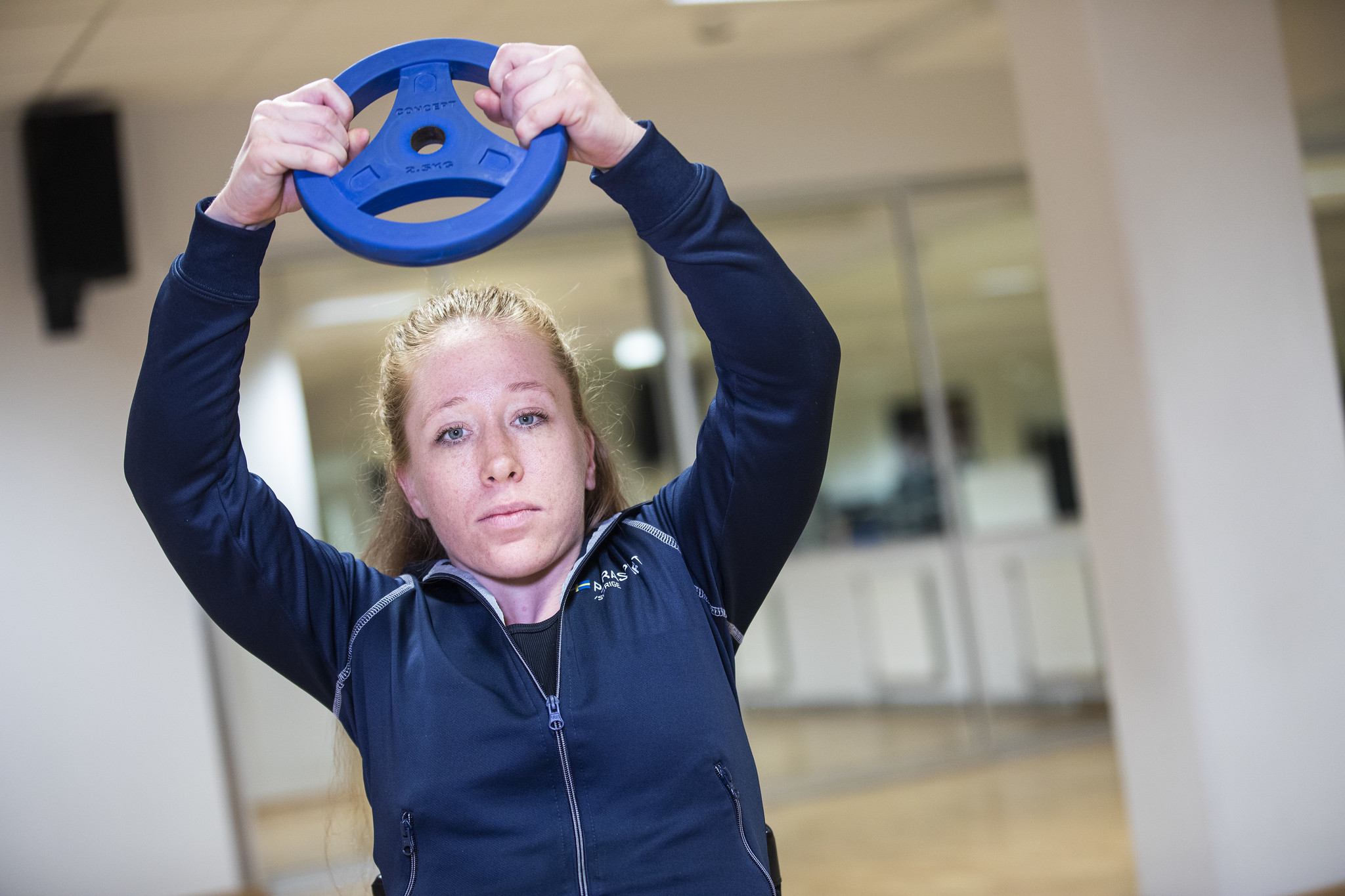 På bilden syns en kvinna med mörkblå, långärmad tröja och uppsatt hår. Hon håller en blå, rund vikt ovanför huvudet med båda händerna. Bakgrunden är suddig men en pelare och några speglar är synliga.