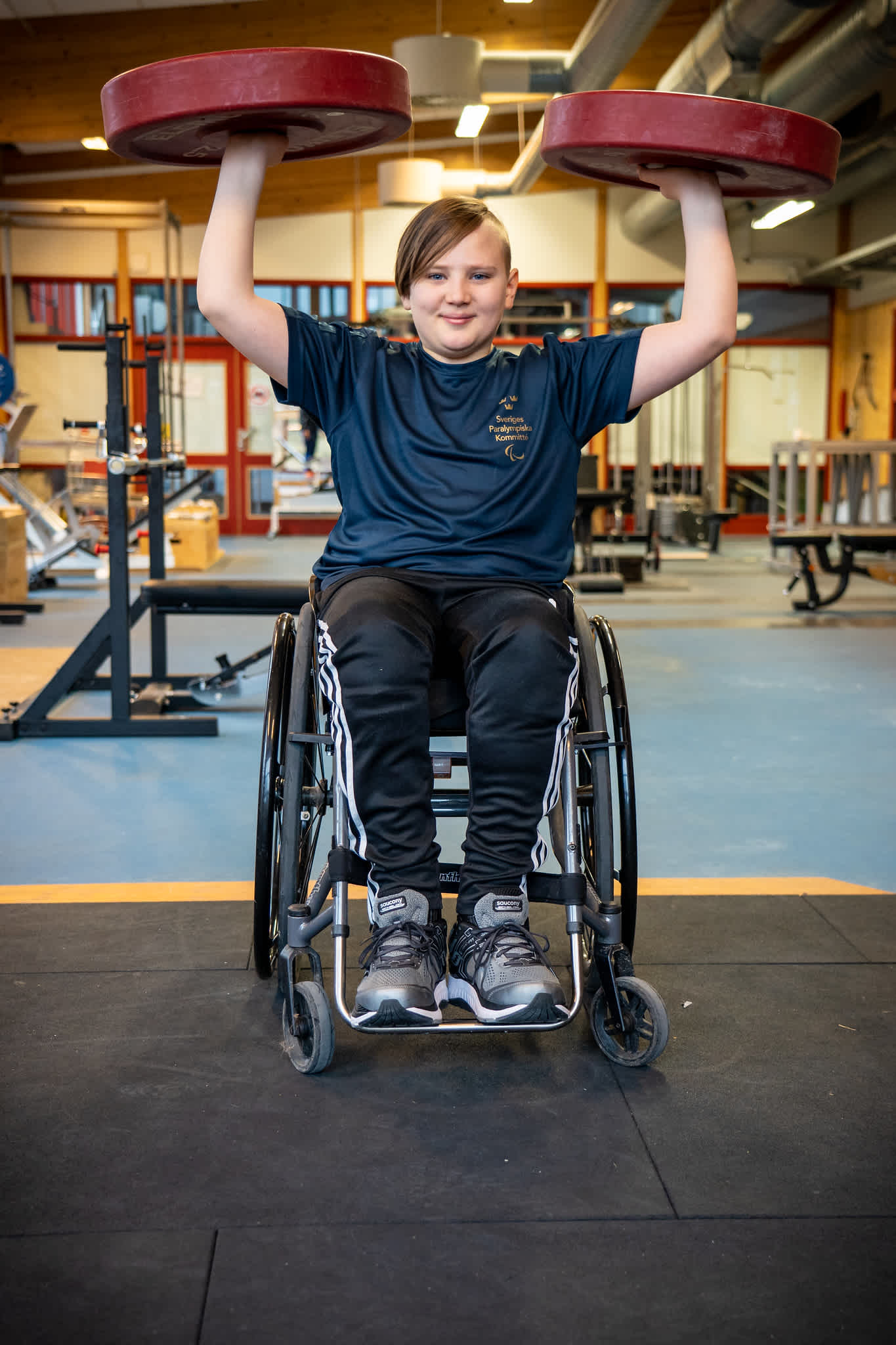 Ung kille sitter i rullstol och håller upp två skivstångsvikter med armarna.