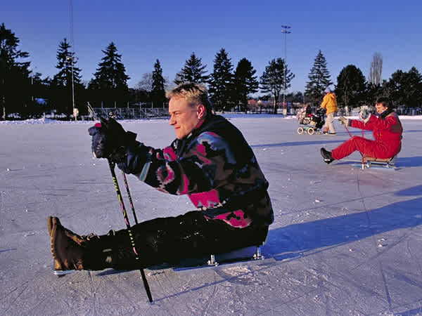 Utomhusmiljö, isrink. En man i skridskokälke stakar sig fram med stavar. I bakgrunden en annan åkare. Foto: Spinalis, Tomas Engblom.