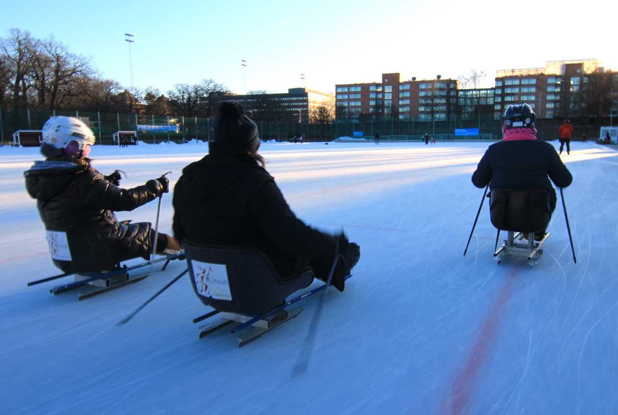 Utomhusmiljö, isrink. Tre skridskokälkeåkare åker på en isrink, i bakgrunden blå himmel och flervåningshus. 