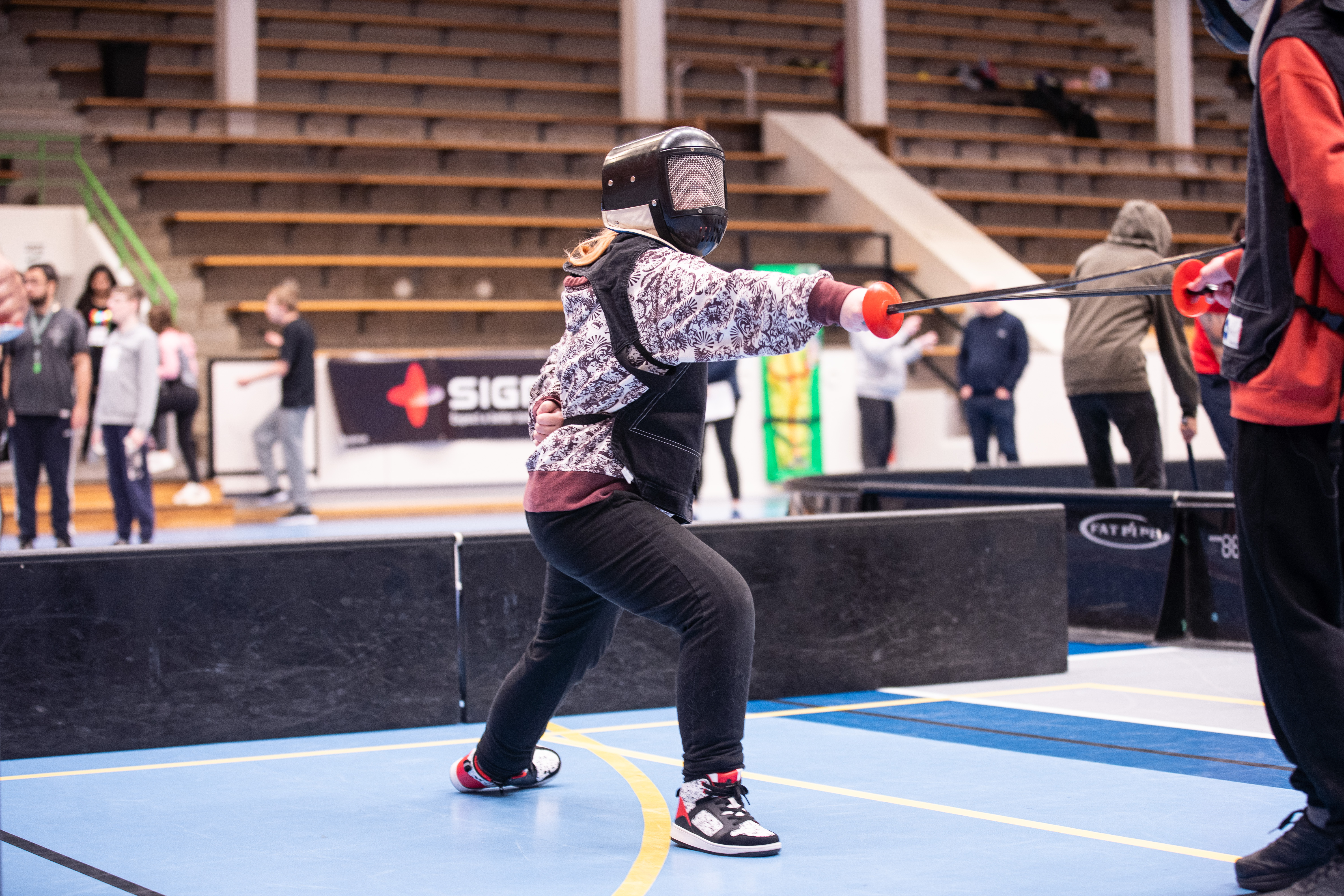 En ung person fäktas, precis gjort ett utfall med höger ben, i höger hand har hon värjan och armen är rak och riktat mot motståndaren, lätt böjt knä och vänster hand bakom ryggen.