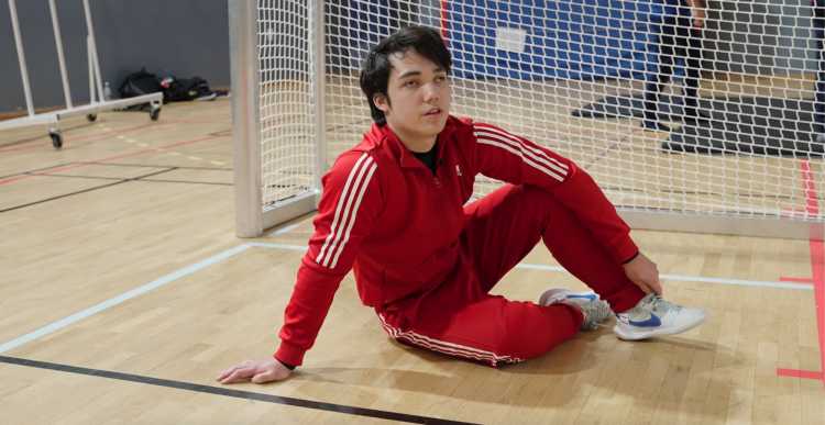Inomhusmiljö, idrottshall. En man i röd träningsoverall sitter på golvet framför att mål. Han har mörkt hår och gympaskor.