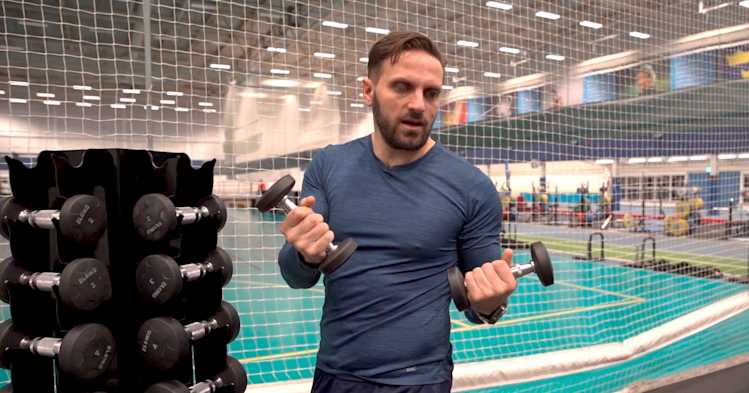 Inomhusmiljö, gym. En man håller upp två hantlar, han står bredvid en hantelställning. I bakgrunden är det en träningsyta.