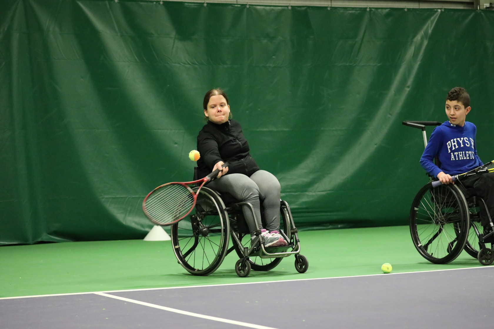 En tjej slår till tennisbollen. En kompis sitter bredvid och kollar på.