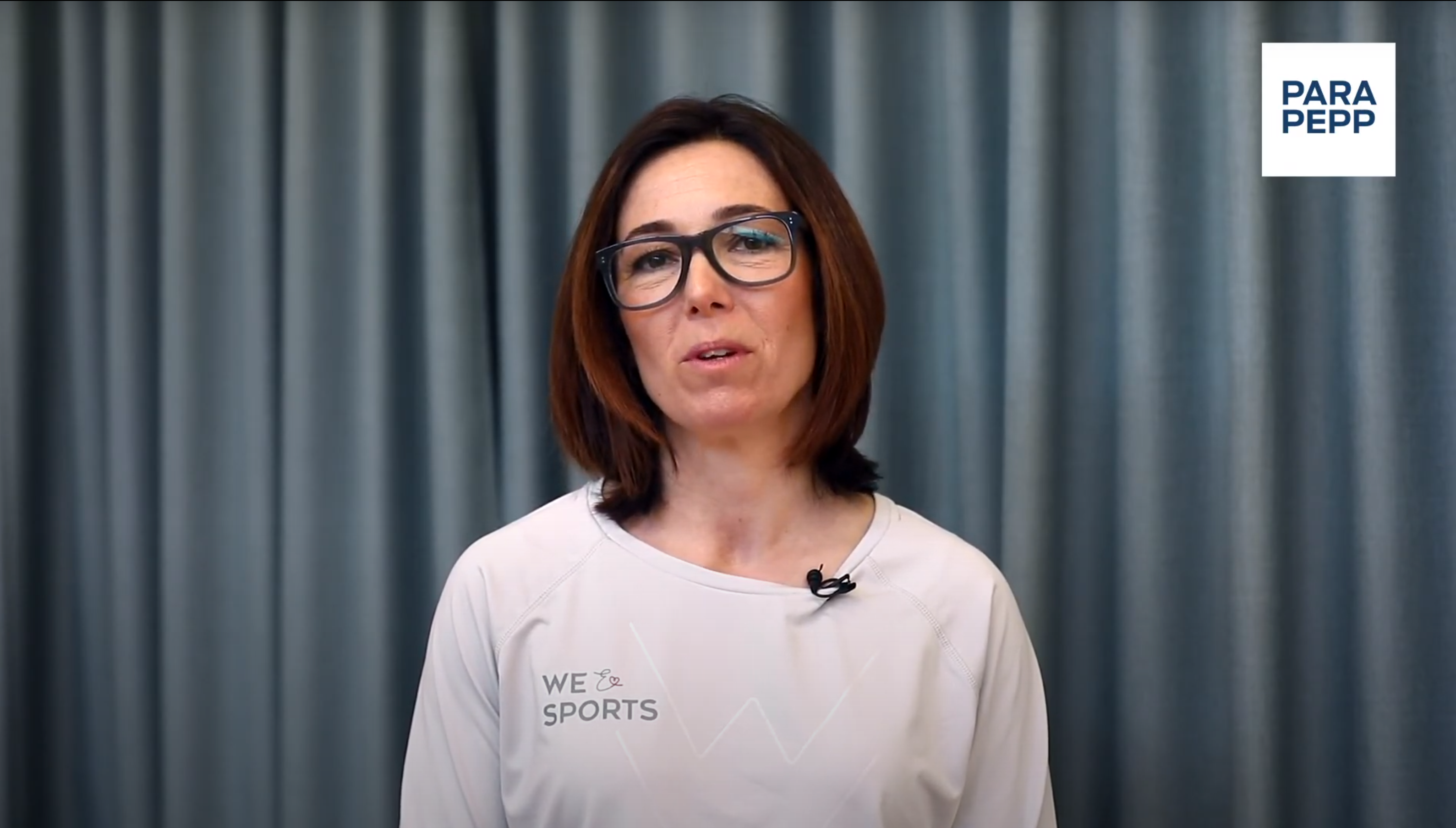 En kvinna med mörkbrunt, axellångt hår och svarta glasögon tittar in i kameran. Hon har en långärmad, vit tröja på sig och bakgrunden är grå. 